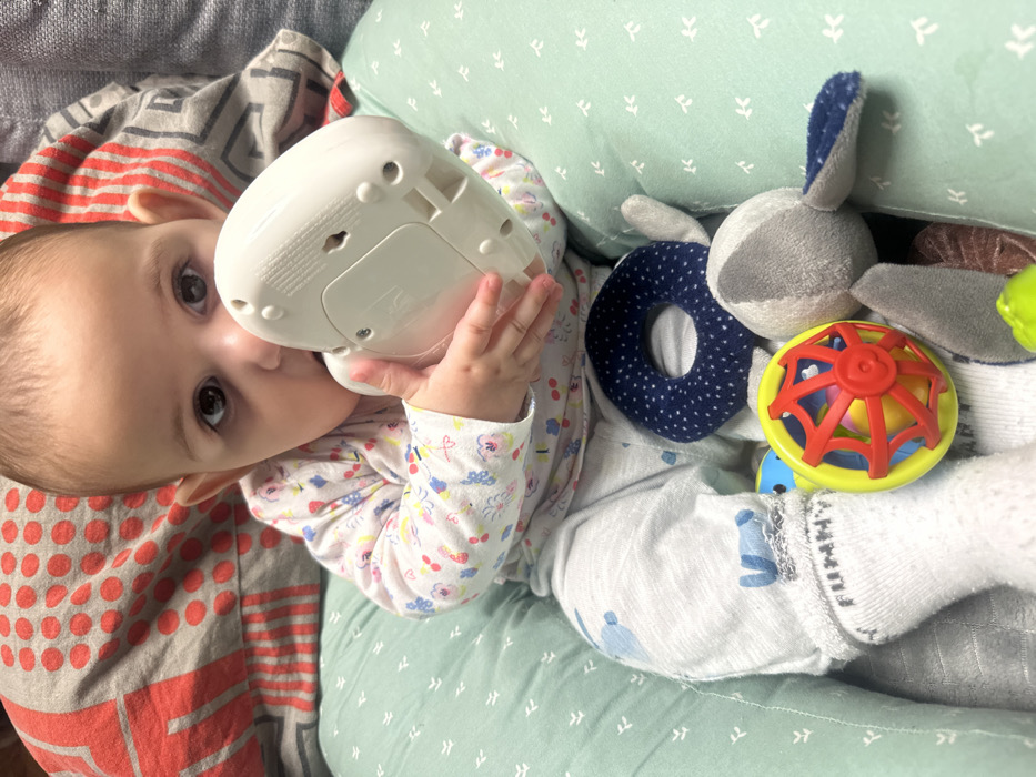 A baby lies on a green pillow with white patterns, clutching a white electronic device. The baby wears a patterned onesie and white socks. To the right, a navy donut-shaped toy, a gray rabbit plush, and a colorful ball with red and yellow elements are visible. Part of a patterned blanket with gray and red stripes is on the left. The baby's gaze is directed at the viewer.