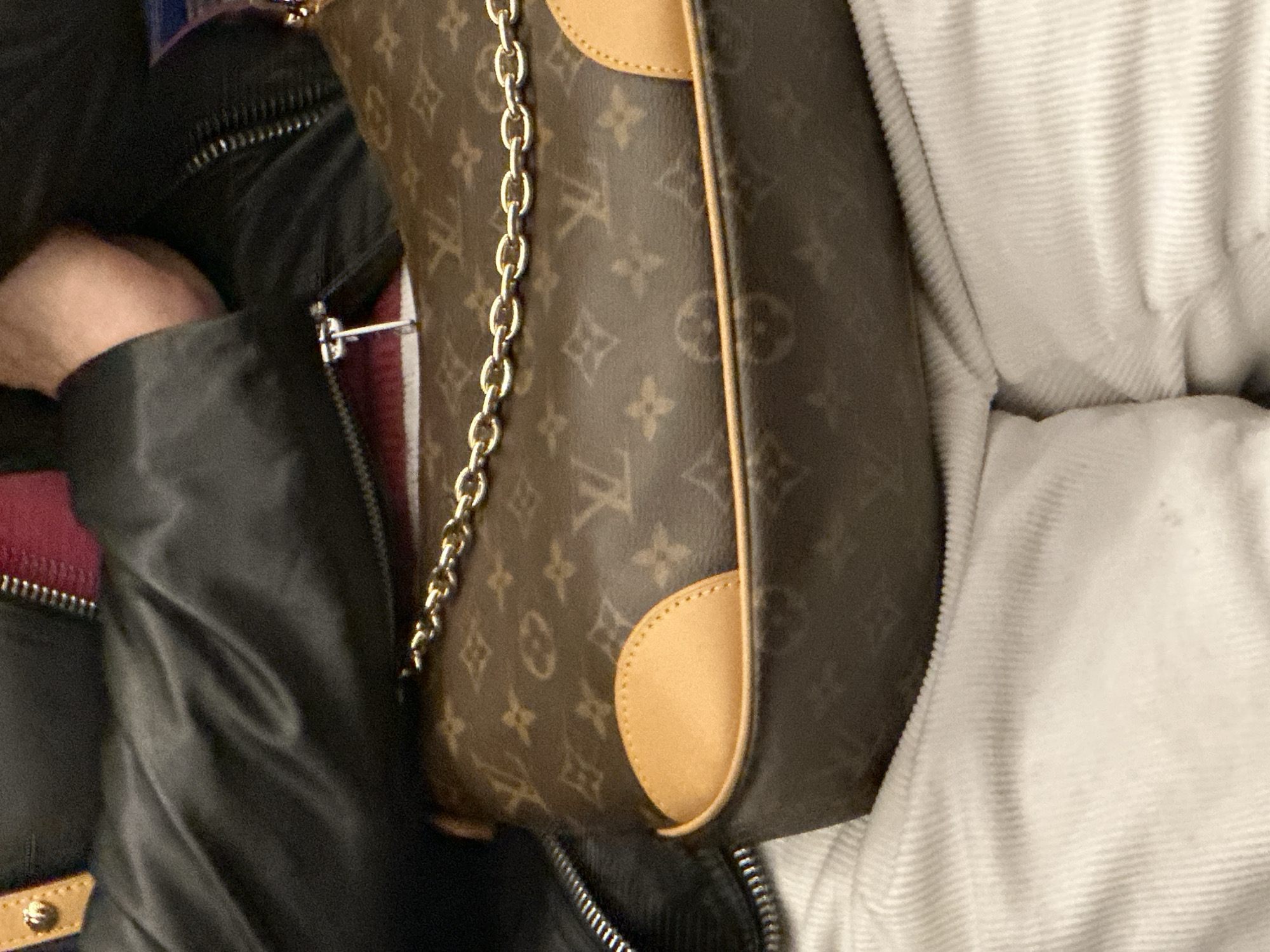 The image presents a close-up view of a person and a handbag, captured in what is understood to be London, United Kingdom. On the left, a person's arm is visible, encased in a black jacket made of a somewhat shiny fabric, possibly nylon. A prominent silver zipper runs down the front of this jacket, and a glimpse of a reddish garment or lining with another zipper is seen beneath it. The person's hand is partially visible within the jacket sleeve opening.
The central subject is a brown handbag, clearly identifiable by its Louis Vuitton monogram pattern. The pattern consists of repeating "LV" initials, quatrefoils, and stylized flowers in a lighter brown tone against a darker brown background. A gold-toned chain strap is draped over the top portion of the bag. The bag features light tan leather trim, particularly noticeable on a rounded, stitched patch on its side.
To the right of the bag, a light-colored fabric, appearing white or off-white, with a distinct ribbed or corduroy texture, occupies the remainder of the frame. This material could be another piece of clothing worn by the person or upholstery. The scene appears to be indoors or in a confined space, with no specific time of day or weather conditions discernible. No explicit text is visible apart from the recurring Louis Vuitton monogram. The overall impression is a casual, unposed moment.