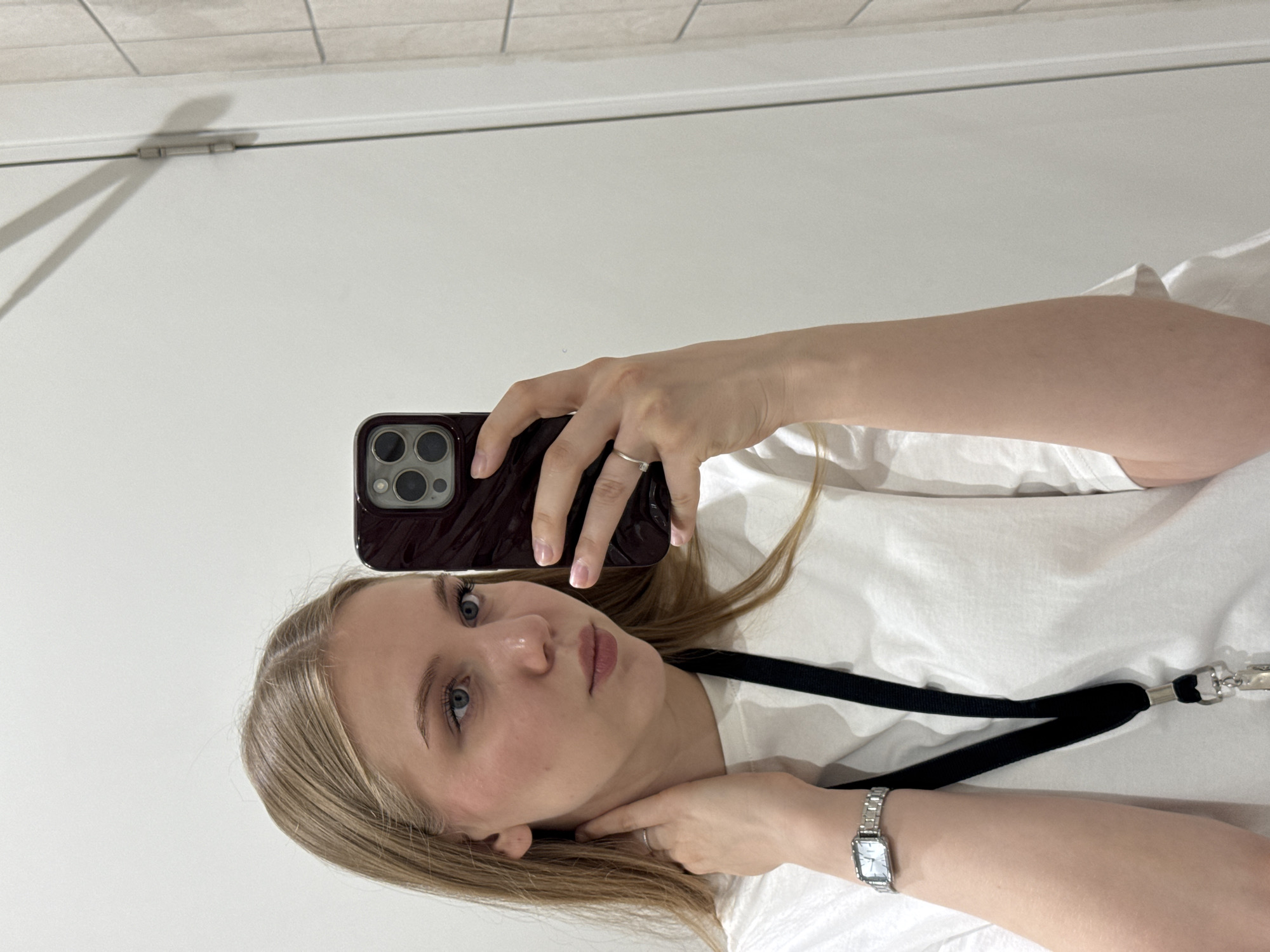 A young female, appearing to be in her late teens or early twenties, is depicted in a mirror selfie taken indoors in Perm, Russia. She has light blonde hair parted down the middle, blue eyes, and fair skin with a slight flush on her cheeks. Her gaze is directed towards the smartphone she holds in her left hand (viewer's right), which has a dark burgundy case and three prominent rear camera lenses. Her right hand (viewer's left) is positioned with her fingers gently touching her upper neck and collarbone area.
She is wearing a plain white crew-neck t-shirt. A black fabric lanyard with a silver-toned metal clasp and clip hangs around her neck, visible over her shirt. On her right wrist, she wears a silver-toned rectangular watch with a metal link bracelet. A plain silver ring is present on the ring finger of her right hand, and another similar silver ring is visible on the ring finger of her left hand.
The background consists primarily of a plain white wall or door. On the upper right side of the image, a white door is visible, featuring a single silver hinge. To the far right edge of the frame, a section of light-colored, possibly rectangular, wall tiles is discernible.