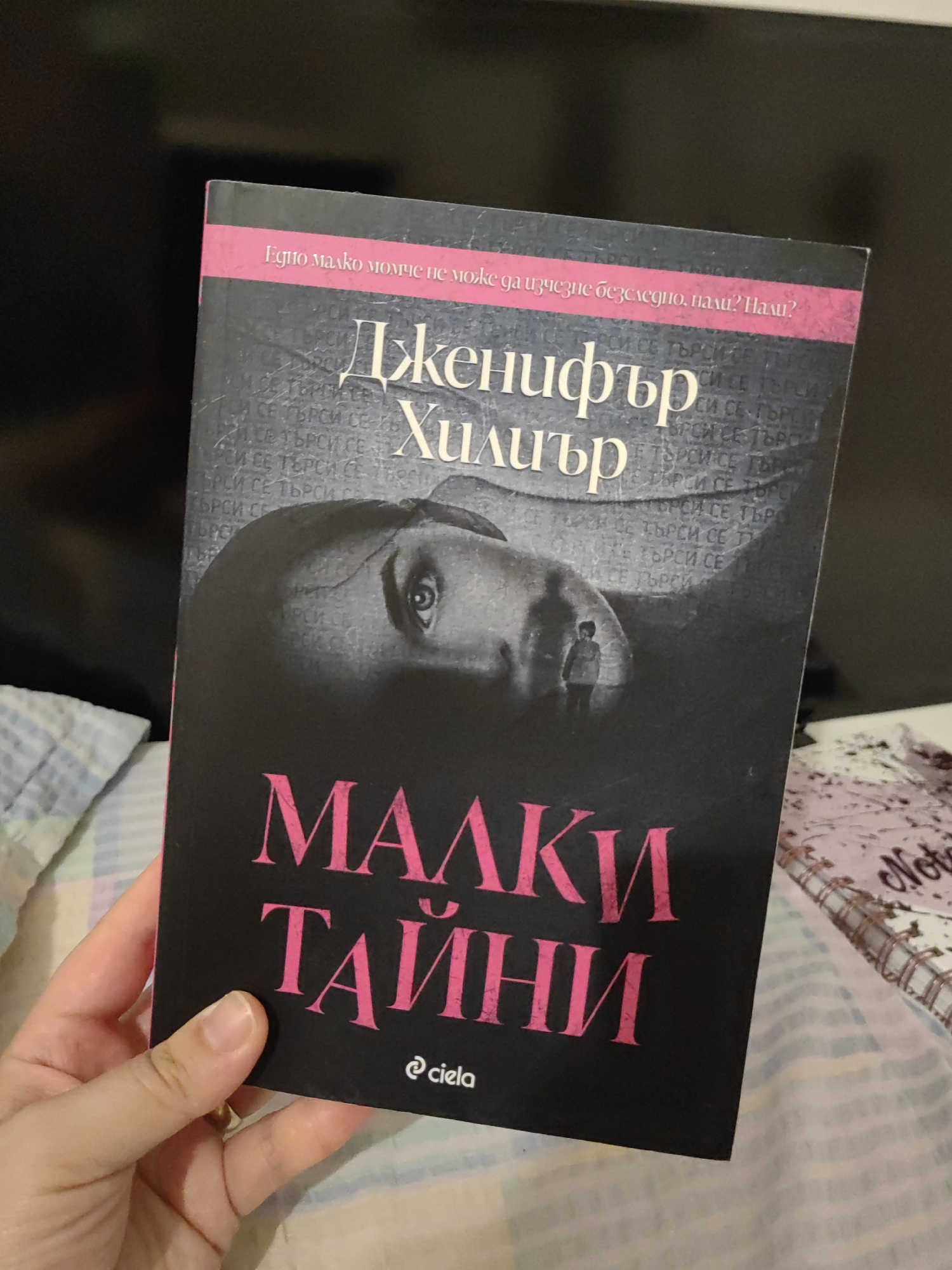 A close-up shot shows a person's hand holding a book titled "Малки тайни" (Malki tayni, meaning "Little Secrets") by Jennifer Hillier. The book cover features a dark, atmospheric image of a woman's eye peering out from shadows, with a silhouetted child figure in the background. The book's title and author's name are prominently displayed in bold, pink lettering. The background appears to be a bed with a patterned blanket. The book is partially illuminated, highlighting the details of the cover. The scene is devoid of any specific landmarks or contextual clues that would indicate a location within Djerovo, Bulgaria.