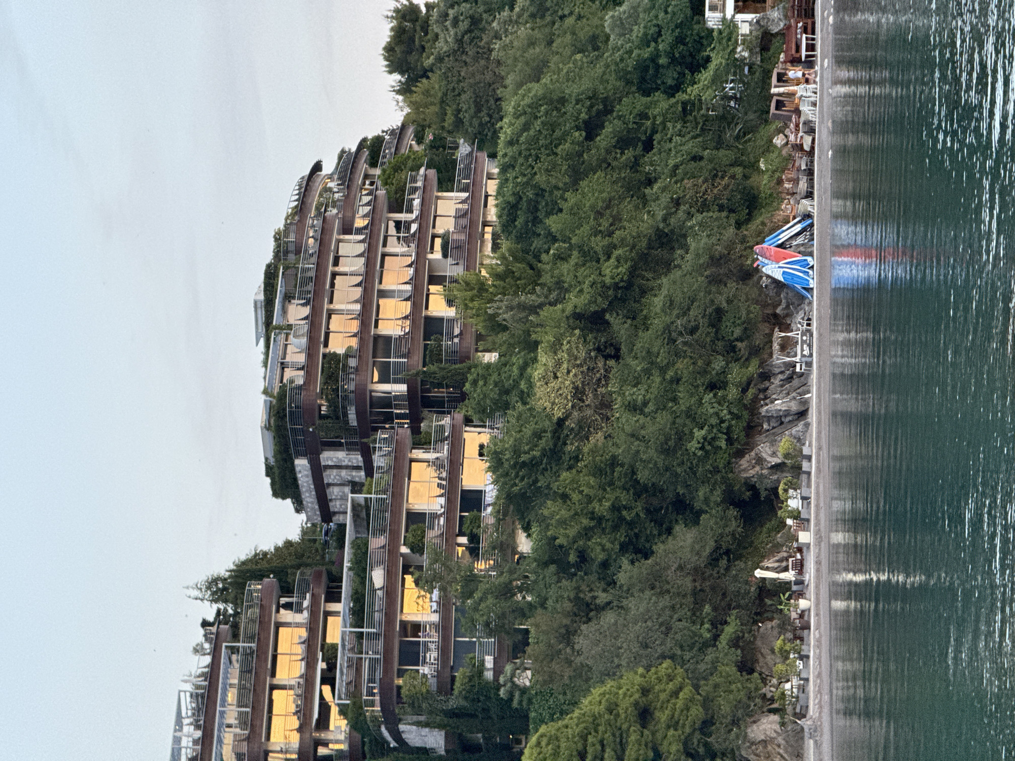 The image captures a tiered, modern hotel complex built into a forested hillside overlooking a body of water. The hotel has a curved, multi-level design with numerous balconies and large windows reflecting the warm light of what appears to be sunset or sunrise. Lush green trees surround the hotel and descend down to a small, pebbly beach area. On the beach, several paddleboards are stacked upright, two of which are brightly colored red and blue. There are also lounge chairs, tables, and umbrellas set up on the beach, suggesting a resort or private recreational area. A person is visible walking near the beach structures. The water in the foreground is calm and reflects the light and the surrounding environment. The scene appears to be located in Budva, Montenegro, a coastal town known for its scenic beauty and resort destinations.
