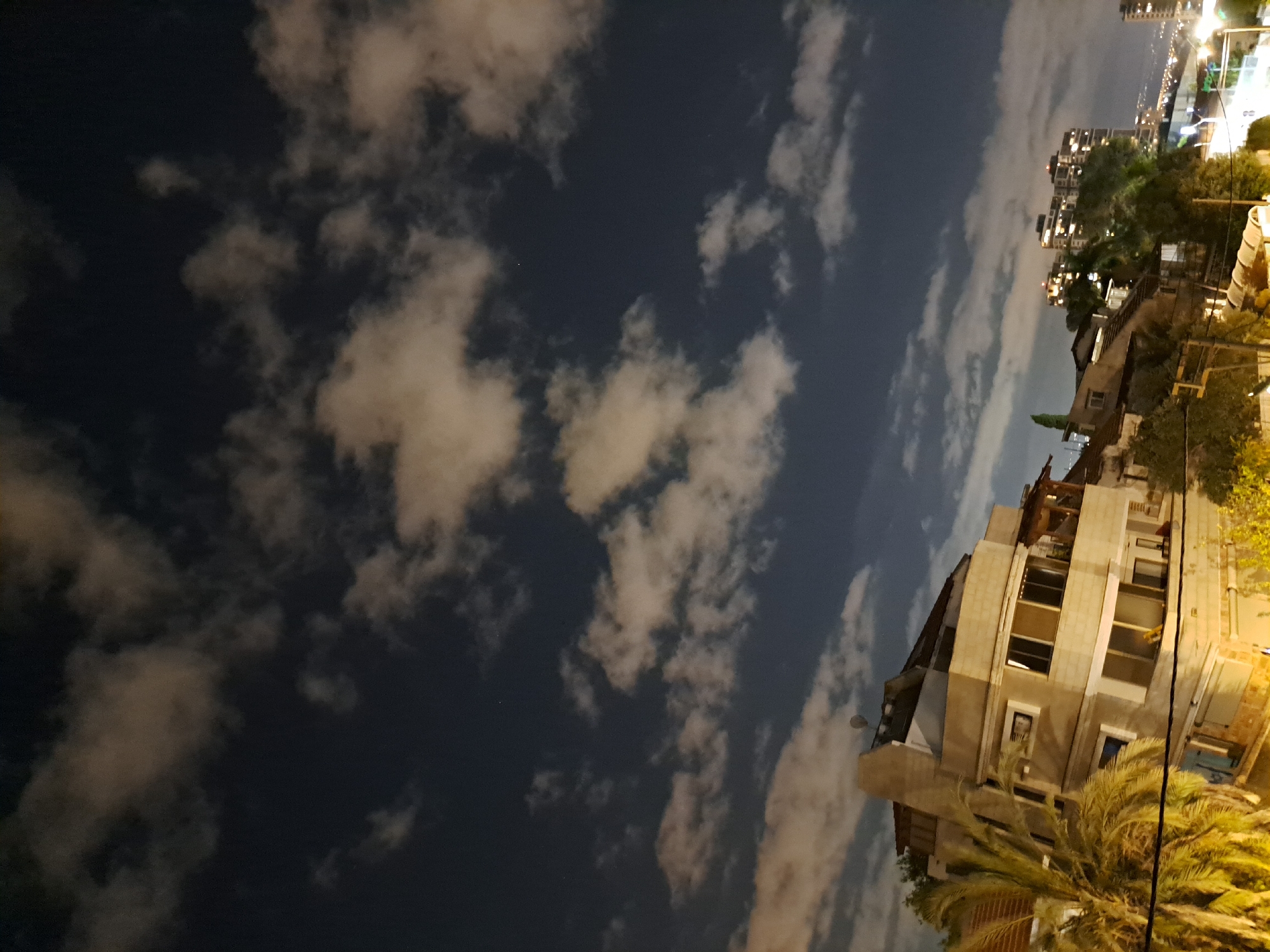 This nighttime, slightly angled, wide shot captures a residential area in Ramat HaSharon, Israel, under a cloudy sky. The foreground features the upper floors of a multi-story building with a light-colored facade, several windows, and balconies. A large palm tree with illuminated fronds partially obscures the left side of the building. In the midground, more low-rise buildings with red-tiled roofs and a few scattered trees are visible. Further in the distance, a tall, modern apartment building, illuminated with many lights, stands out against the dark sky. The horizon shows a more distant city skyline with more lights. The sky is a deep blue-black, with scattered, illuminated clouds creating a dramatic effect. There are no visible people in the frame.