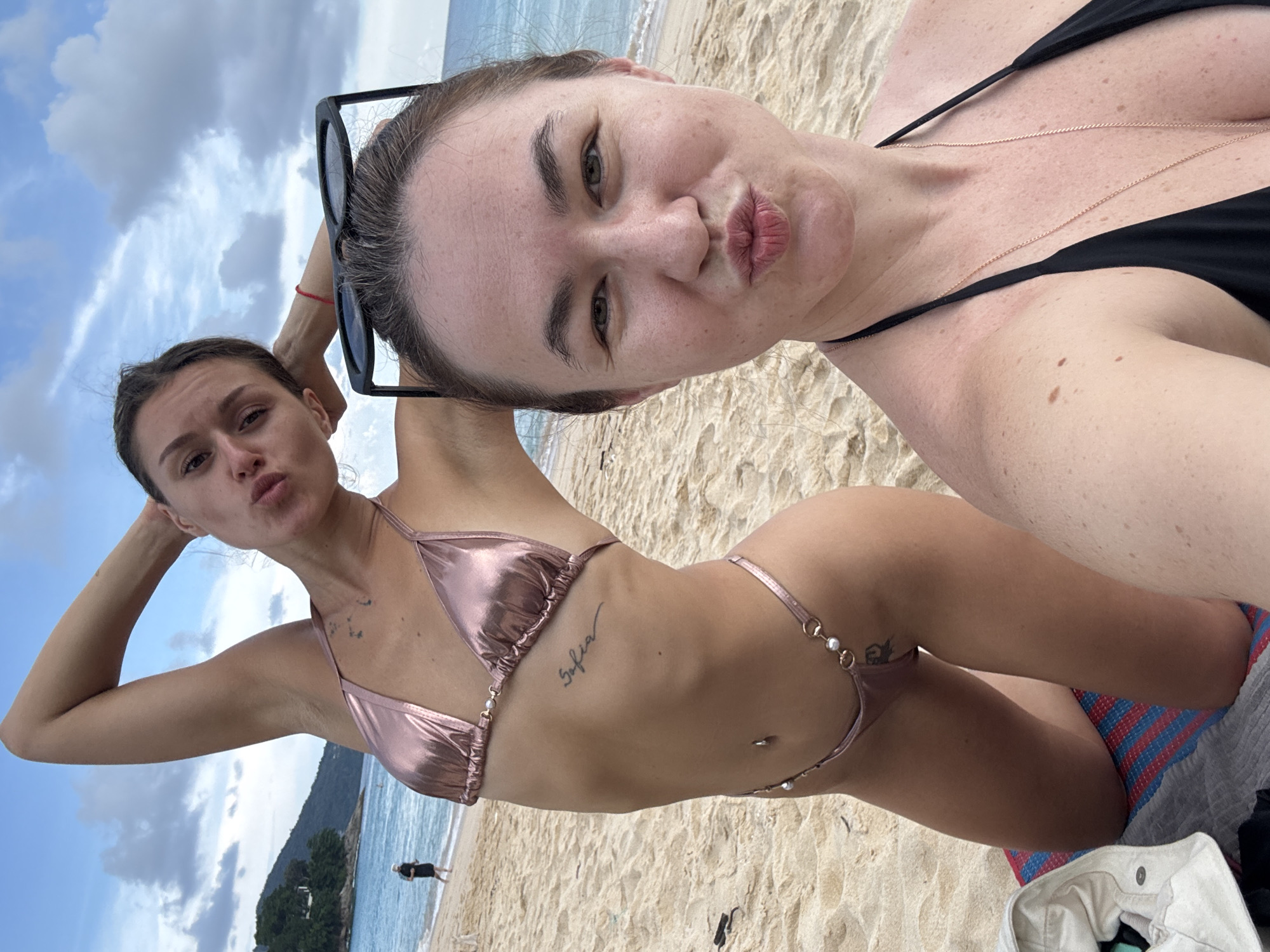Two women are posing for a selfie on a beach in Choeng Thale, Thailand. The woman in the foreground, closer to the camera, is wearing a black bikini and has sunglasses perched on her head. She is puckering her lips for the photo. The woman in the background, who is kneeling on a striped mat, is wearing a metallic pink bikini. She has her arm raised behind her head and is also puckering her lips. The background shows a sandy beach, the ocean, and a cloudy sky. A person is visible walking in the distance near the water.