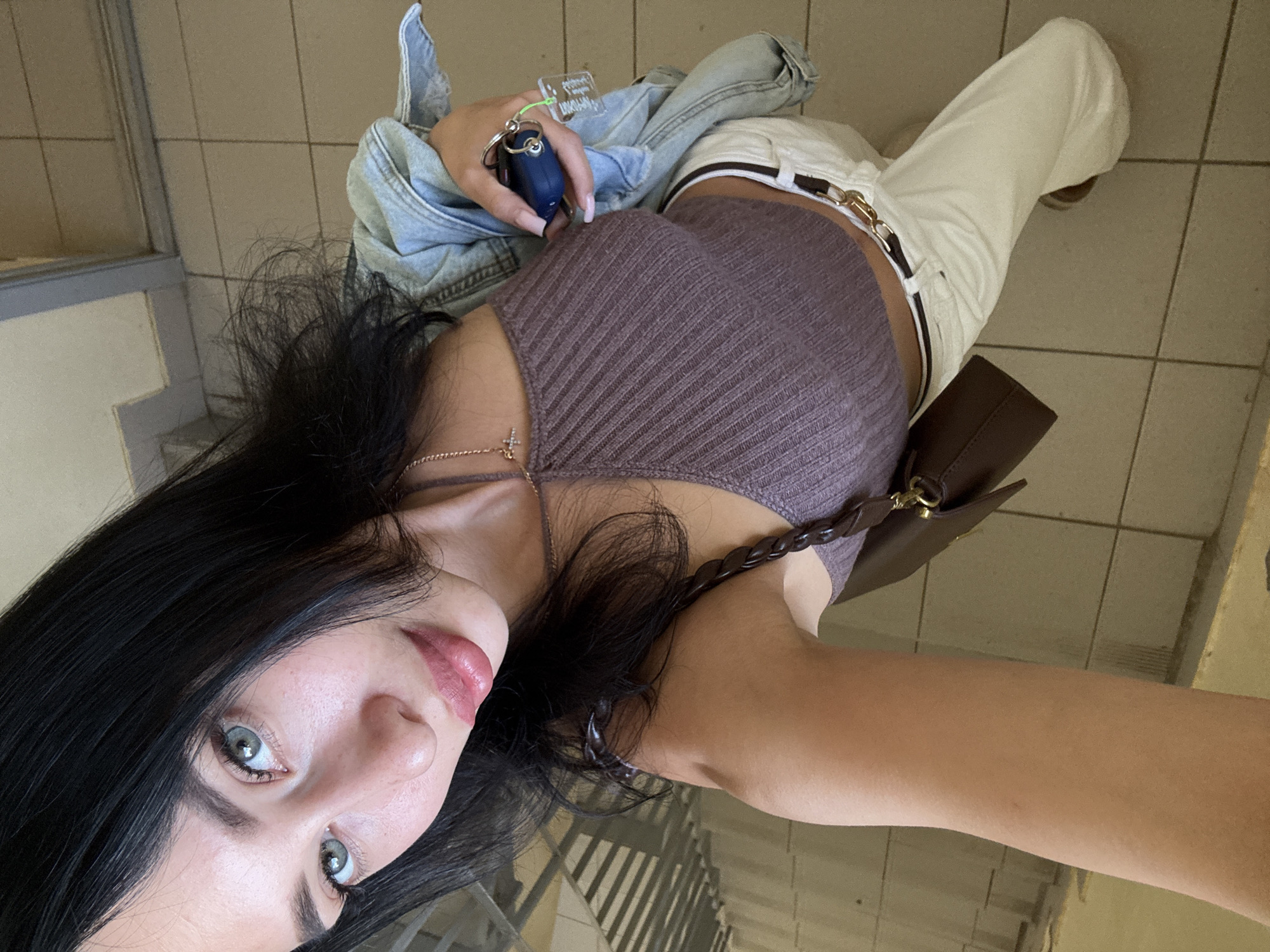 The media file shows a selfie of a young woman from a high angle, looking upwards at the camera. She has long, dark hair and light-colored eyes with visible eyelashes. Her lips are painted with a light pink lipstick. She is wearing a ribbed, dark mauve halter top and cream-colored pants with a white belt and gold buckle. A brown handbag with a braided strap is slung over her shoulder. She is holding a blue car key fob with a clear tag attached. A light blue denim jacket is draped over her left shoulder. The background appears to be an indoor tiled floor and wall, suggesting a public or indoor space. The lighting is bright, indicating it's daytime. No specific weather conditions are discernible, but the overall impression is casual and everyday.