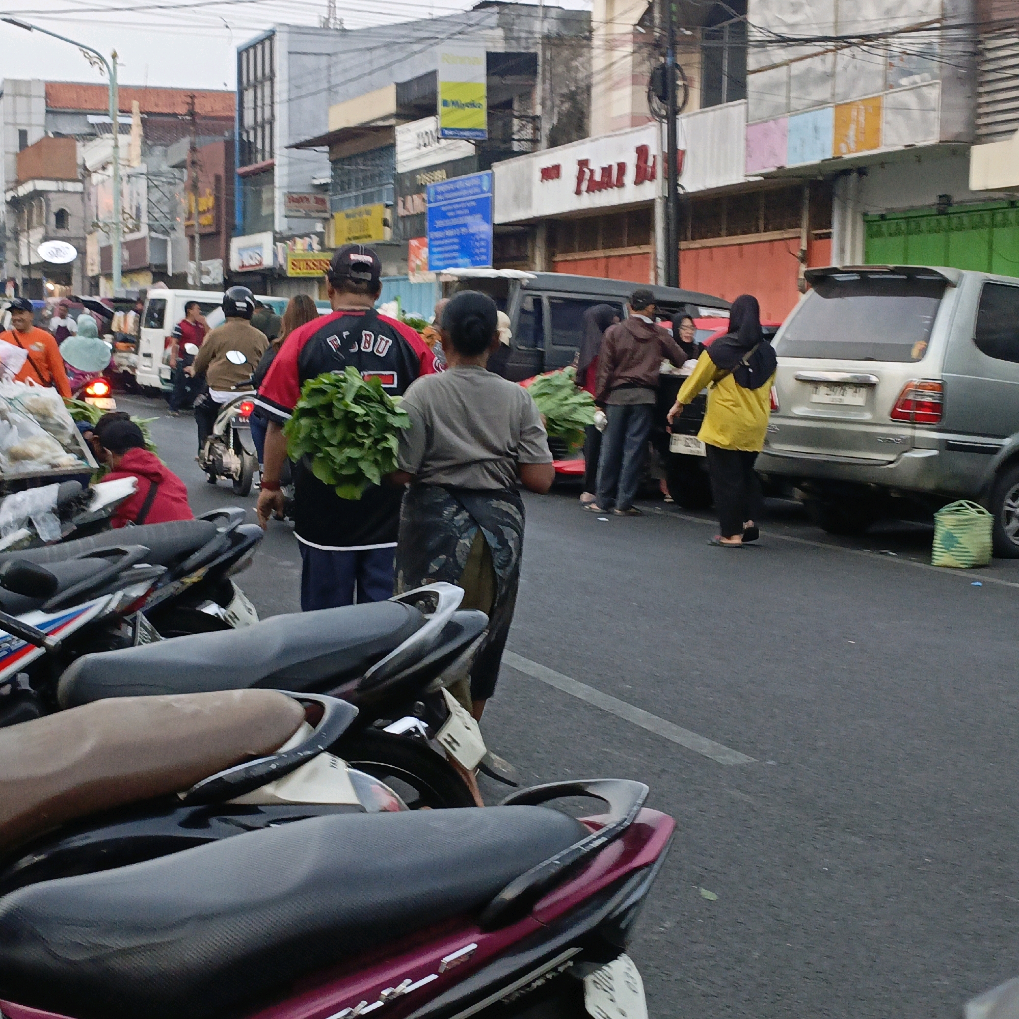 This is a street scene in Salatiga, Indonesia, likely a market area. In the foreground, several motorcycles are parked, with their seats visible. A person wearing a black and red shirt with white lettering and a black baseball cap is holding a large bunch of green leafy vegetables. Next to them, another person, possibly a woman, is wearing a grey t-shirt and a patterned skirt or sarong, also carrying a bunch of leafy greens. Further back, a person in a yellow jacket and black headscarf walks away from the camera, alongside a silver SUV. Other people and vehicles, including a white van and a motorcycle with a rider, are visible in the background. Buildings line the street, some with signs. The overall impression is a busy, everyday scene in an Indonesian town.