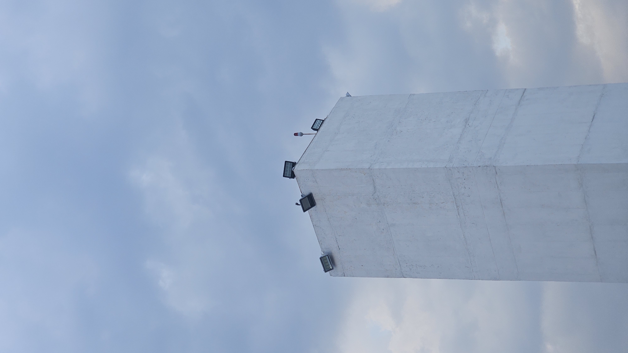 A low-angle shot shows a tall, white, possibly concrete structure against a cloudy sky. The structure appears to be a tower or pillar, and it is adorned with several rectangular spotlights or floodlights mounted near the top. An additional element is visible at the top of the tower, possibly a red safety beacon. The background is filled with a mix of light blue and white clouds, suggesting an overcast day. The image is likely from San Bernardino Tlaxcalancingo, Mexico, given the instruction to consider that location.