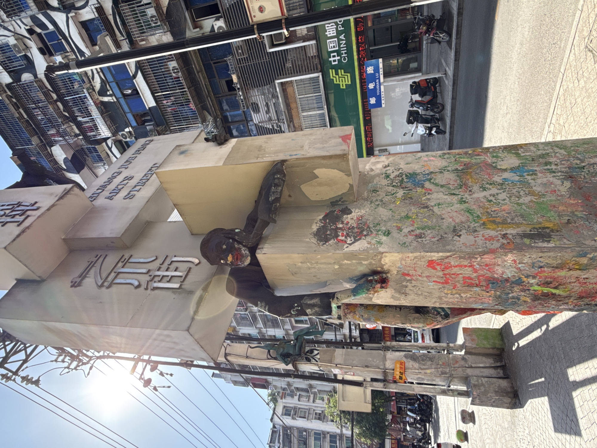 The image shows a street scene in Jiulongpo District, China, featuring the entrance to Huang Jue Ping Arts Street. The entrance is marked by a large, cross-shaped monument or sign, painted in grey tones and covered in colorful paint splatters. A dark-colored statue of a figure appears to be integrated into the monument. The street name "Huang Jue Ping Arts Street" is written in both English and Chinese characters on the monument. Buildings of varied heights and architectural styles line the street. Chinese signage for "China Post" and a street name "Youdian Lu" are visible. Scooters and motorcycles are parked along the street, while overhead power lines crisscross the sky.