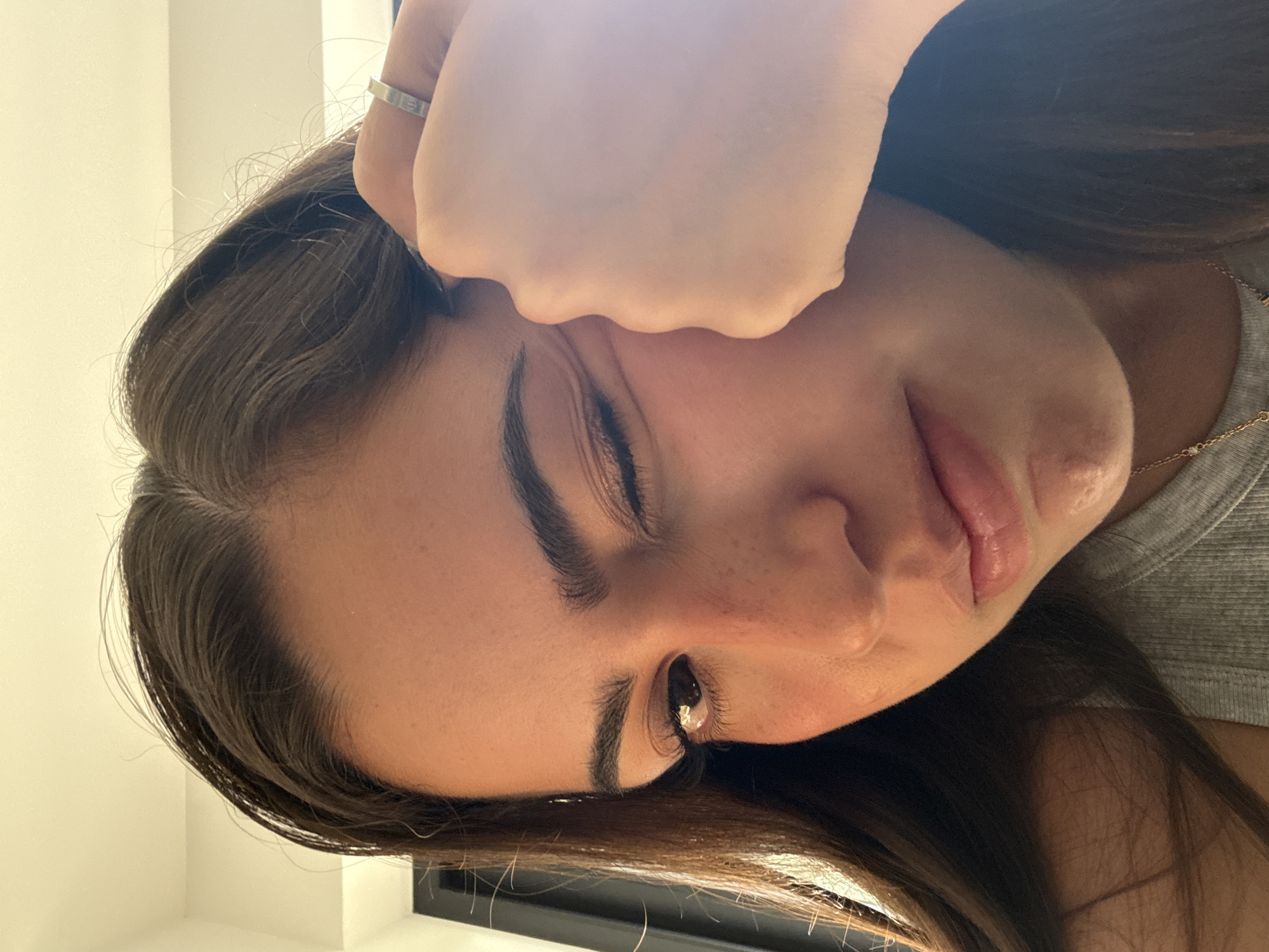 A close-up, slightly tilted portrait of a woman with brown hair, light skin, and dark eyebrows and eyelashes. Her left eye is open, revealing a brown iris, while her right eye is closed. Her left hand, adorned with a silver ring, is positioned against her forehead. She is wearing a gray top and a delicate gold necklace. The background consists of a white wall in the upper left corner and a dark shape suggesting a window frame or object in the lower portion of the image.
There is no verifiable information to ascertain this image being taken in San Bernardino Tlaxcalancingo, Mexico.