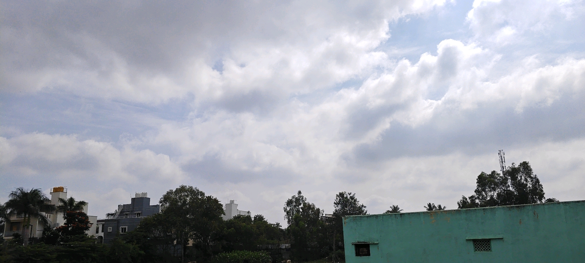 This is a panoramic, eye-level, outdoor, daylight photograph taken in Hennagara, India. The image depicts a cloudy sky with patches of blue visible. In the foreground, the top of a teal-colored building with a small, screened window is visible. Beyond the building, a dense line of green trees forms a backdrop. Amongst the trees, several multi-story residential buildings of varying architectural styles and colors, including a white building with a yellow water tank and a dark grey building, can be seen. A cell tower is also visible above the trees towards the right side of the frame. No people or animals are present in the scene. The overall impression is a residential area on a partly cloudy day.