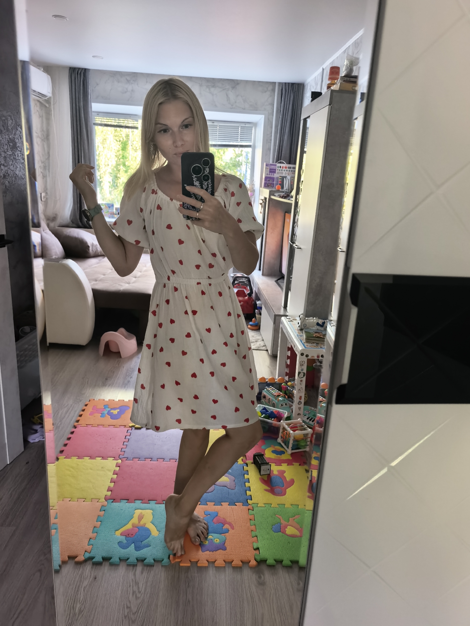 A woman is taking a mirror selfie in what appears to be a living room in Sterlitamak, Russia. She is wearing a white dress with red hearts and is holding her phone to take the picture. The room has a large mirror, a couch, and a window with a view of trees. The floor is covered with colorful foam mats. There are also toys and furniture visible in the background.