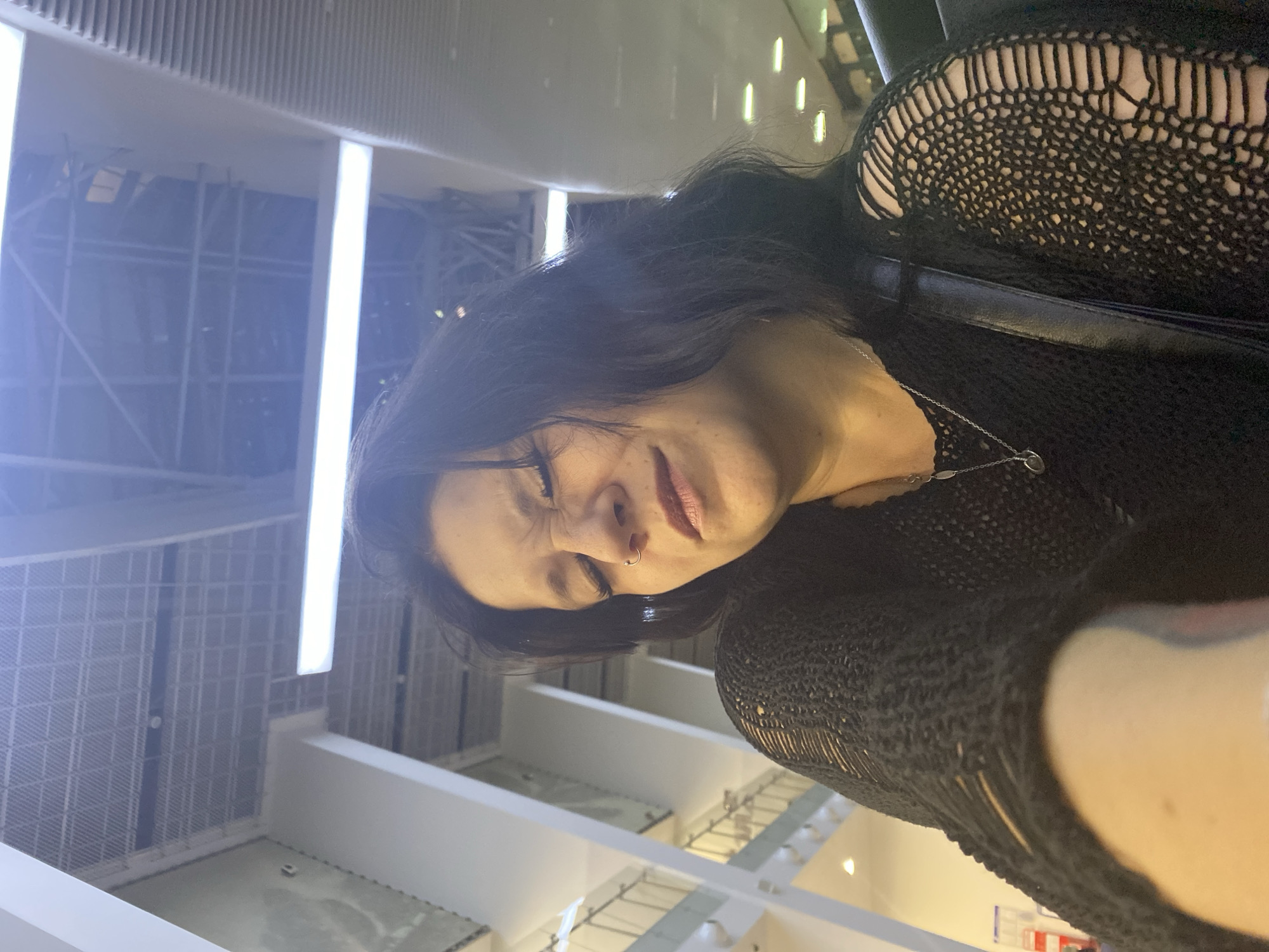 The image shows a woman with dark hair and a nose ring, wearing a black knitted top. Her eyes are closed and her facial expression suggests discomfort or pain. She appears to be indoors, in a large open space with a high ceiling and visible lighting fixtures and structural elements. The background suggests an interior of a modern building, possibly an airport terminal or a large public space. The specific location in Bolshoye Savino, Russia cannot be determined from the visual information provided in the image.