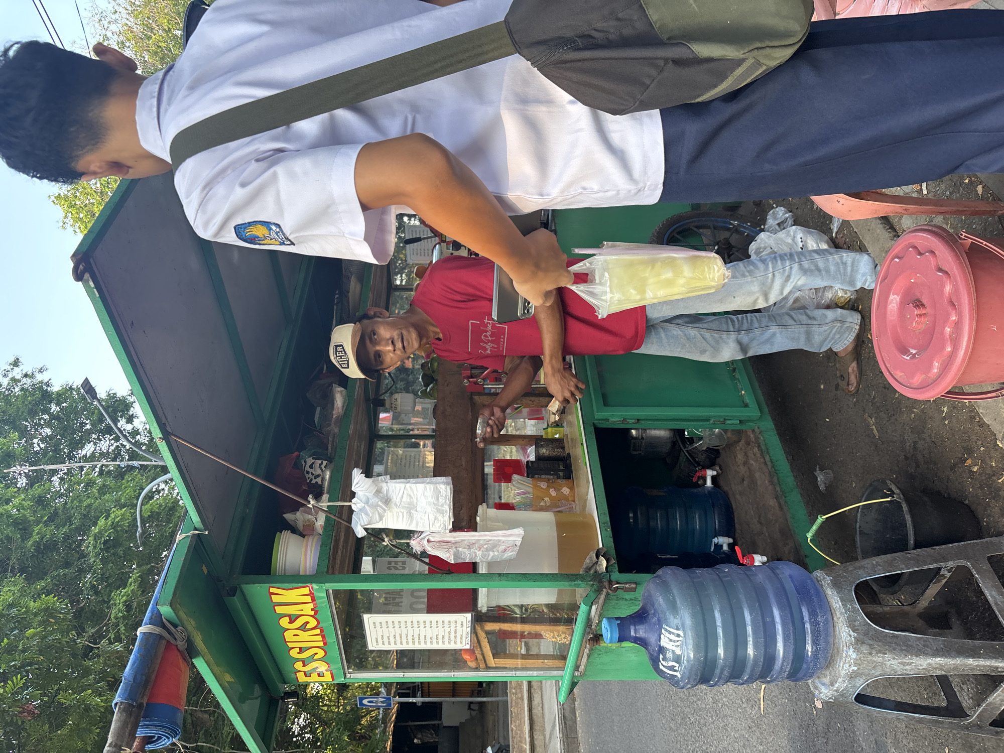 The image depicts a street food vendor in Cirebon, Indonesia, selling drinks from a green cart. A young man in a white school uniform is the customer, receiving a plastic bag with a yellow drink from the vendor. The vendor, wearing a red shirt and a beige cap, appears to be in the process of serving the customer. Behind the vendor is a large container of a yellowish liquid, presumably the drink being sold. The cart is decorated with the words "ES SIRSAK" in large yellow letters, indicating it sells soursop juice. Several large water jugs are visible next to the cart, along with buckets and other supplies. The scene takes place outdoors, with trees and a street in the background, suggesting a public area in the city. The lighting indicates it is likely daytime.