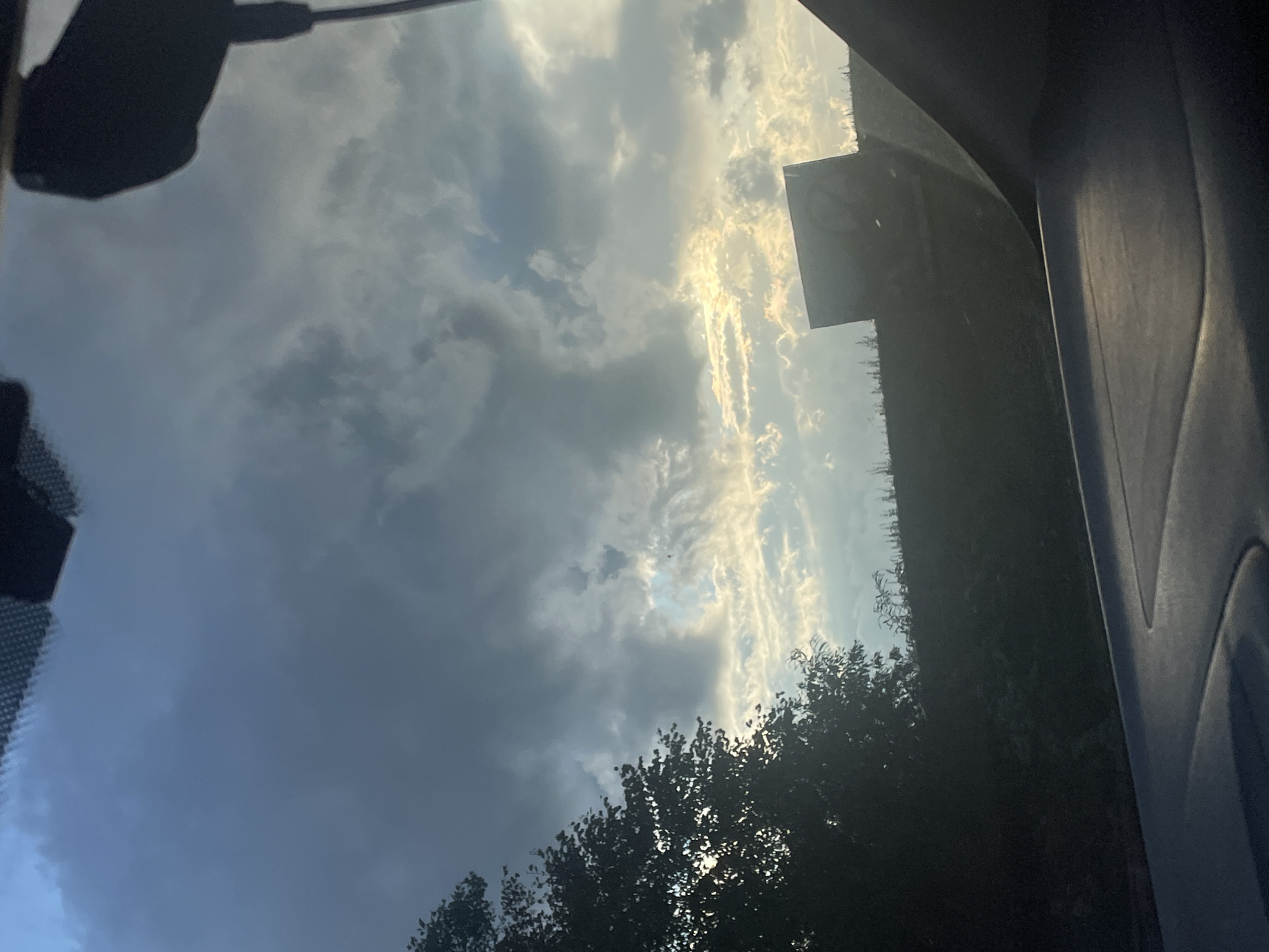 The image is an outdoor shot, taken from inside a vehicle looking upwards. The scene depicts a dramatic sky with a mix of dark, ominous clouds and bright, sunlit streaks of clouds, suggesting a dynamic weather condition. The sun appears to be low in the sky, possibly indicating either sunrise or sunset. Silhouetted against the bright sky are the tops of trees and a portion of a dark, angular object, possibly a building or a signpost. The interior of the car is partially visible, with dark plastic trim on the right side and a textured surface on the left, likely part of the dashboard or windshield. The overall mood is one of nature's grandeur and perhaps a sense of transition, given the interplay of light and shadow in the sky. There is a faint circular symbol with some markings on the square object, but the details are not clear enough to read. The location context is Гавриловка, Russia.
