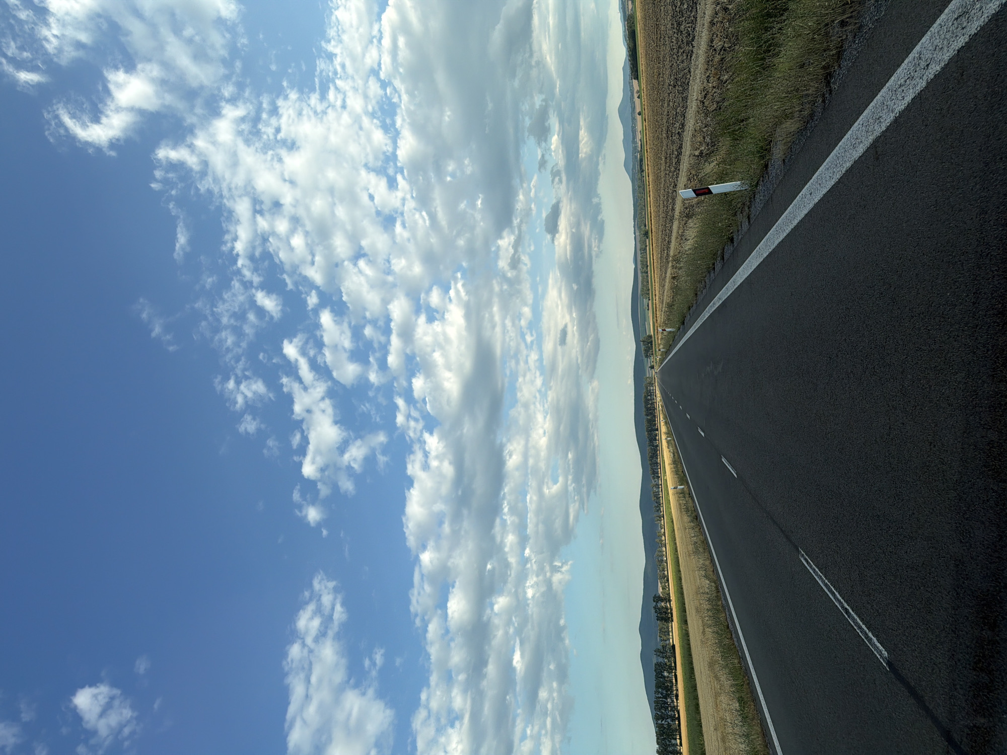 This is a photograph of a road in Kožuchov, Slovakia, with a clear sky filled with white clouds. The road is asphalt and has a white line marking the edge and a double white line in the middle, indicating a two-way street. The road appears to be in good condition.
To the right of the road, there is a field of dry grass and a patch of cultivated land, suggesting it is autumn or a dry season. The cultivated field is a light brown color. There are also a few white road markers with red reflectors on the right side of the road.
To the left of the road, there is a field of golden dry grass and then a line of trees. Further in the distance, there are rolling hills covered in a green forest, and beyond that, mountains are visible under the horizon.
The sky is a vibrant blue with scattered white clouds. Some clouds are large and puffy, while others are thinner and wispy. The lighting suggests it is daytime, possibly late afternoon, as the sun is not directly overhead and the light is somewhat softened.
There are no people or vehicles visible in the image, making it a tranquil landscape scene. The overall impression is one of open space and a journey through the Slovak countryside.