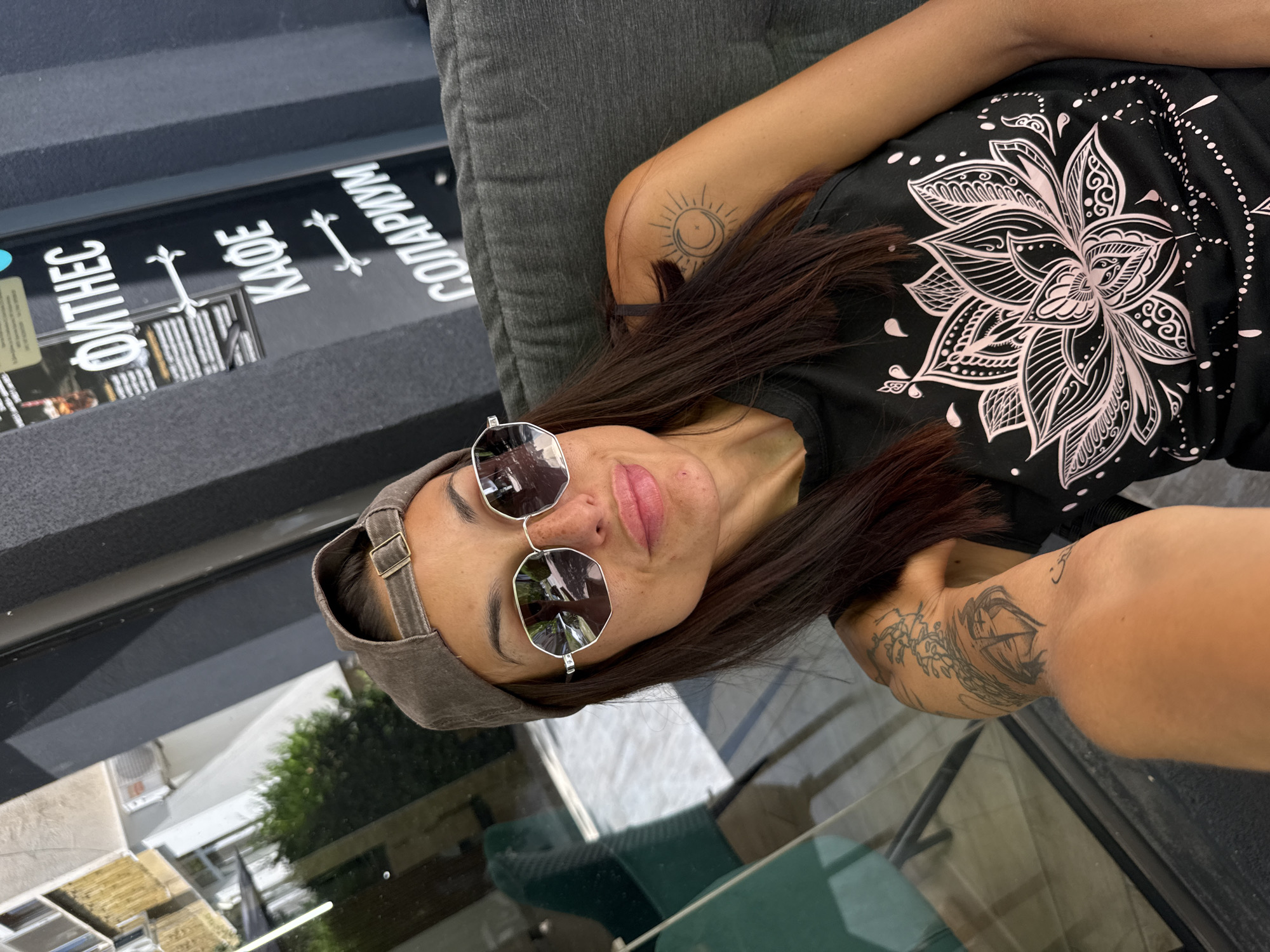 A woman wearing a grey baseball cap and large, polygonal sunglasses is seated on a dark grey sofa, taking a selfie. She has dark hair and is wearing a black tank top with a pink lotus flower design. She has tattoos on her left arm. Behind her, a dark grey building features signage in white lettering, including "ФИТНЕС", "КАФЕ", and "СОЛАРИУМ". The background also shows a reflection of a building and greenery in a glass window. The scene appears to be outdoors, possibly in front of a establishment in Pleven, Bulgaria.