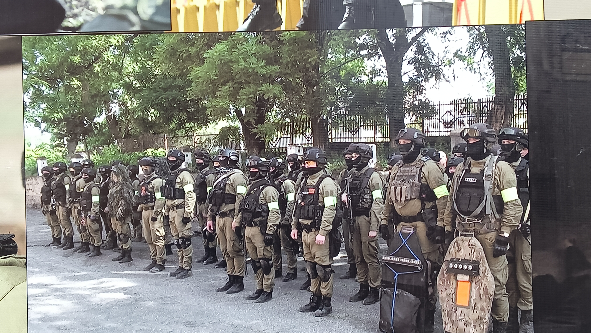 The image displays a group of approximately two dozen individuals, likely military or paramilitary personnel, standing in formation outdoors. They are uniformly equipped in tactical gear, including combat helmets, balaclavas or full face masks, tactical vests (plate carriers), and uniforms predominantly in olive green and tan shades. A notable feature is the bright yellow or light green armbands worn by many of the individuals on one or both sleeves.
In the foreground on the right, two individuals are equipped with large tactical shields. One shield features a camouflage pattern with a prominent orange rectangular marking and circular elements at the top; the other is dark-colored with blue straps. Visible text on the tactical vest of one individual reads "СПЕЦ," possibly indicating "Special Forces" or "Special Purpose."
The scene is set outdoors during daylight hours, with an overcast or partly cloudy sky suggested by the soft lighting. Trees and a dark fence or gate form the background, with the ground being paved. The individuals' posture suggests a formal line-up, potentially for a briefing, inspection, or deployment. The photograph itself appears to be an image captured from a screen. The location is Tiraspol, Moldova.
