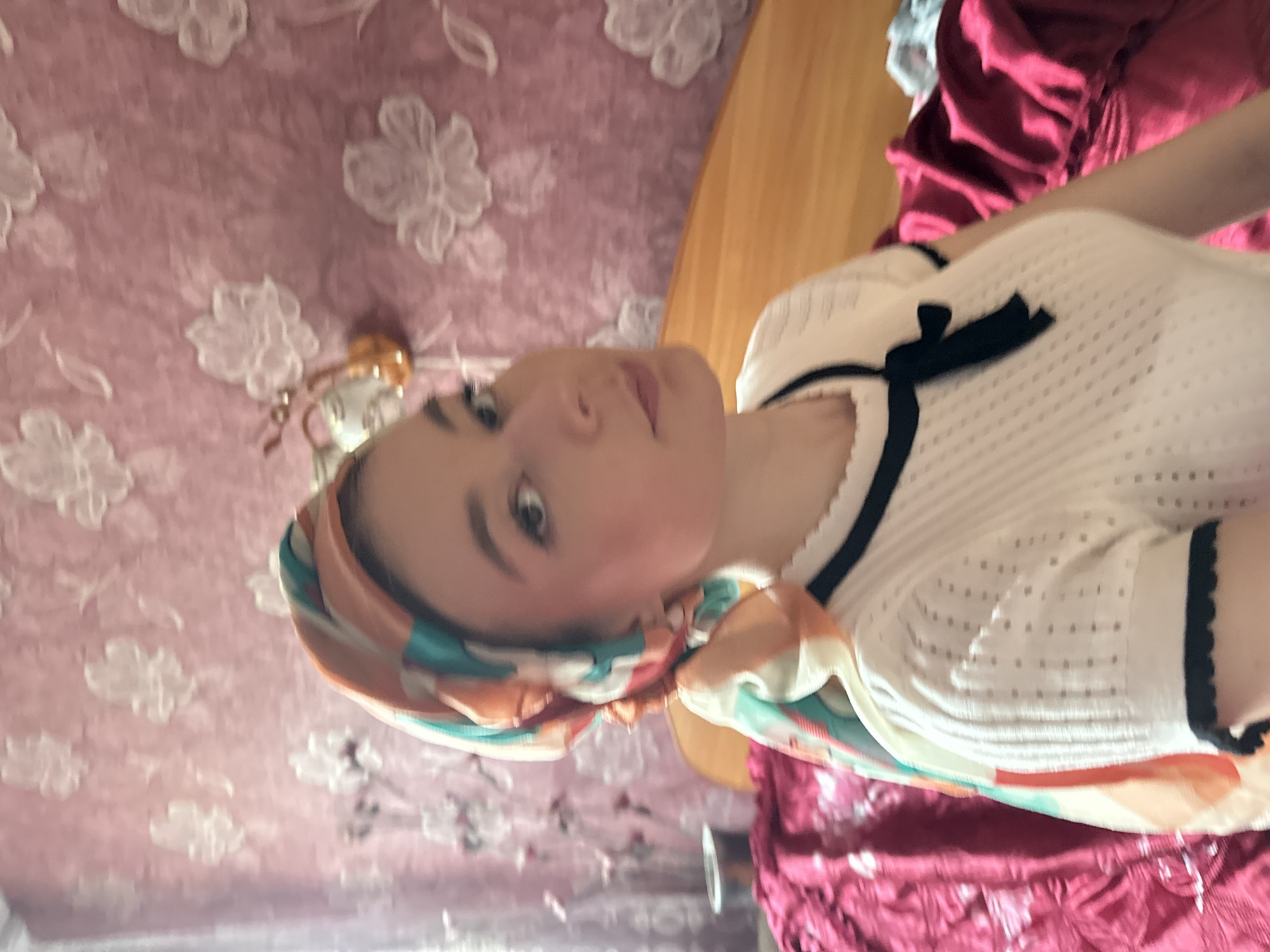 A woman with light skin and brown hair is wearing a headscarf with orange and turquoise patterns. She is wearing a white shirt with black trim and a small black bow at the neckline. The background includes a pink wall with a white floral pattern. It is impossible to ascertain if the photo was taken in Коноваловка, Russia or any specific geographical location.