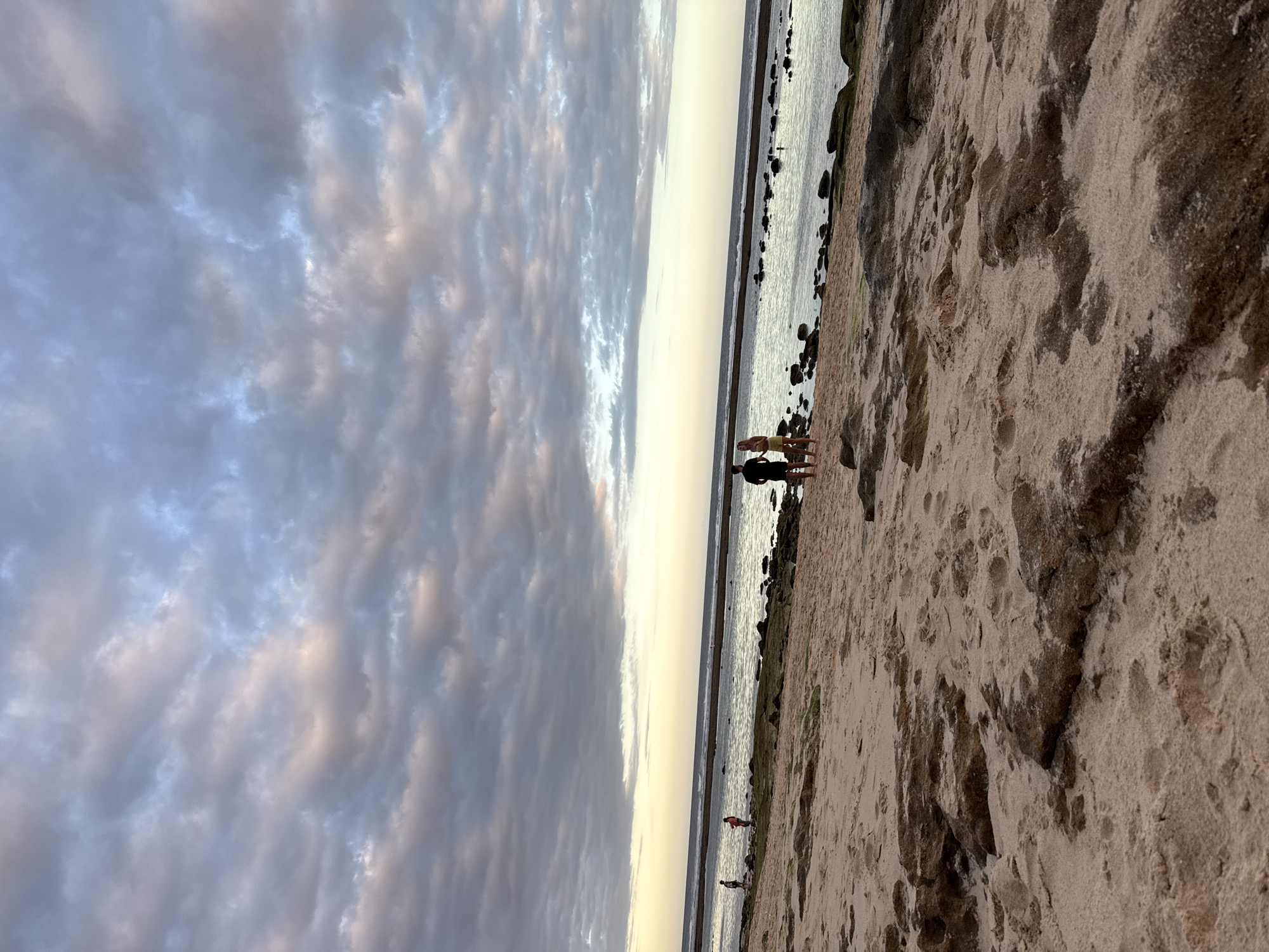 The image depicts a beach scene in Kuta Selatan, Indonesia. The sky is filled with layered clouds, transitioning to a lighter horizon above the ocean. The shoreline consists of sand with some rocks or outcroppings. Several people are present on the beach; some stand near the water's edge, and others are farther down the beach.