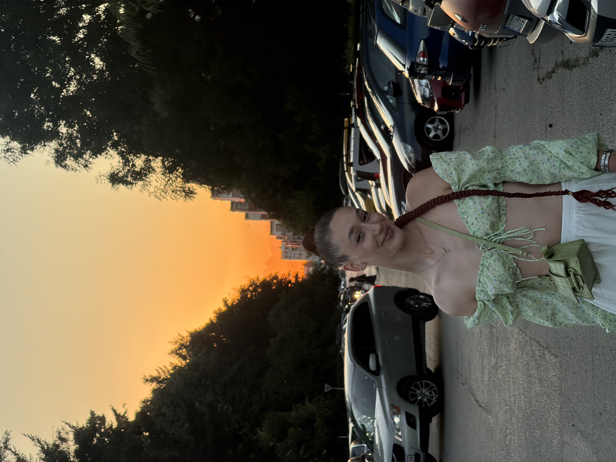 A young woman with her hair in a bun and a long braid stands in a parking lot during sunset in Sofia, Bulgaria. She is wearing a green floral off-the-shoulder crop top and white pants, with a small green handbag slung over her shoulder. The setting sun casts a warm orange glow over the scene, illuminating the trees lining the street and the parked cars. The woman is looking towards the camera with a slight smile. The background shows a street lined with buildings and trees, typical of urban areas in Sofia.