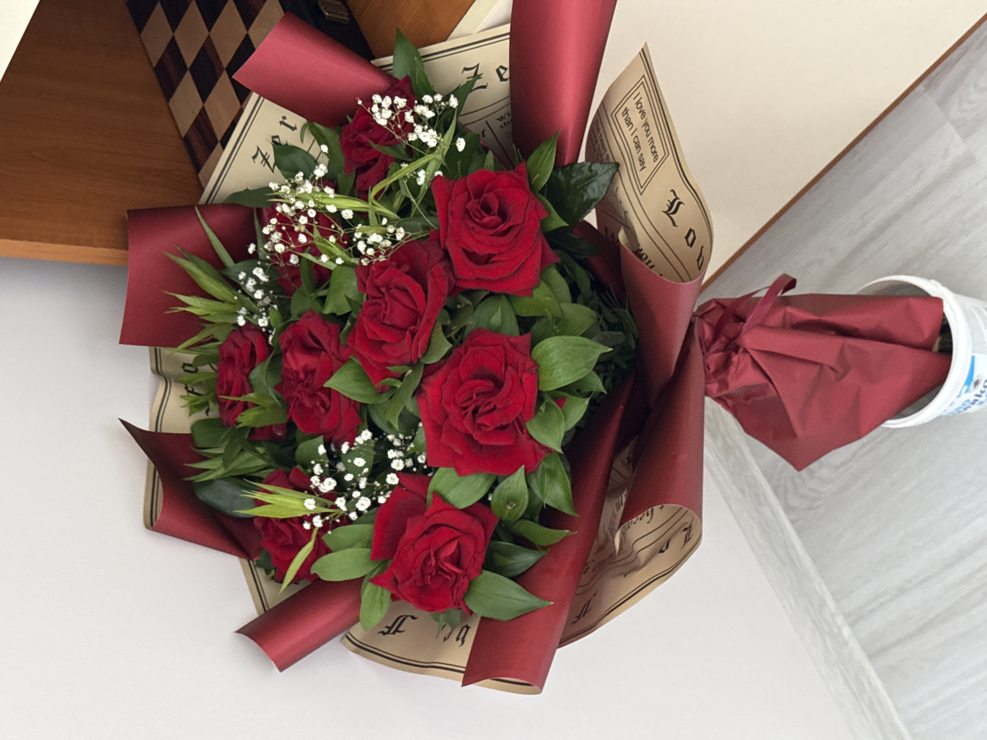 A vibrant bouquet of red roses dominates the center of this image, presented in a high-angle shot. The bouquet features numerous fully bloomed and budding red roses, interspersed with lush green foliage and delicate sprigs of white gypsophila (baby's breath).
The flowers are elegantly wrapped in a two-layered packaging. The outer layer consists of a deep, rich red, slightly glossy paper, artfully folded around the bouquet. The inner layer is made of a lighter brown, rustic-textured paper adorned with text and musical notation. Clearly visible on this brown paper is the phrase "I love you more than I can say," enclosed within a decorative border. Other sections of the brown paper show stylized calligraphic letters and musical staff lines with notes, some partially obscured or upside down.
The bouquet is positioned on a light, possibly white or off-white, horizontal surface. In the upper left background, a wooden shelf or piece of furniture is visible, featuring a distinctive dark brown and light brown checkerboard pattern on its top surface. The background also includes a plain white wall and a light-colored wooden floor or surface. To the right, a white cylindrical object, possibly a cup or container, is partially visible, also wrapped in a matching deep red paper and tied with a ribbon. The scene depicts an indoor setting, likely a home, with soft, even lighting suggesting daytime. No people are present, and no specific activity is taking place beyond the presentation of the bouquet.