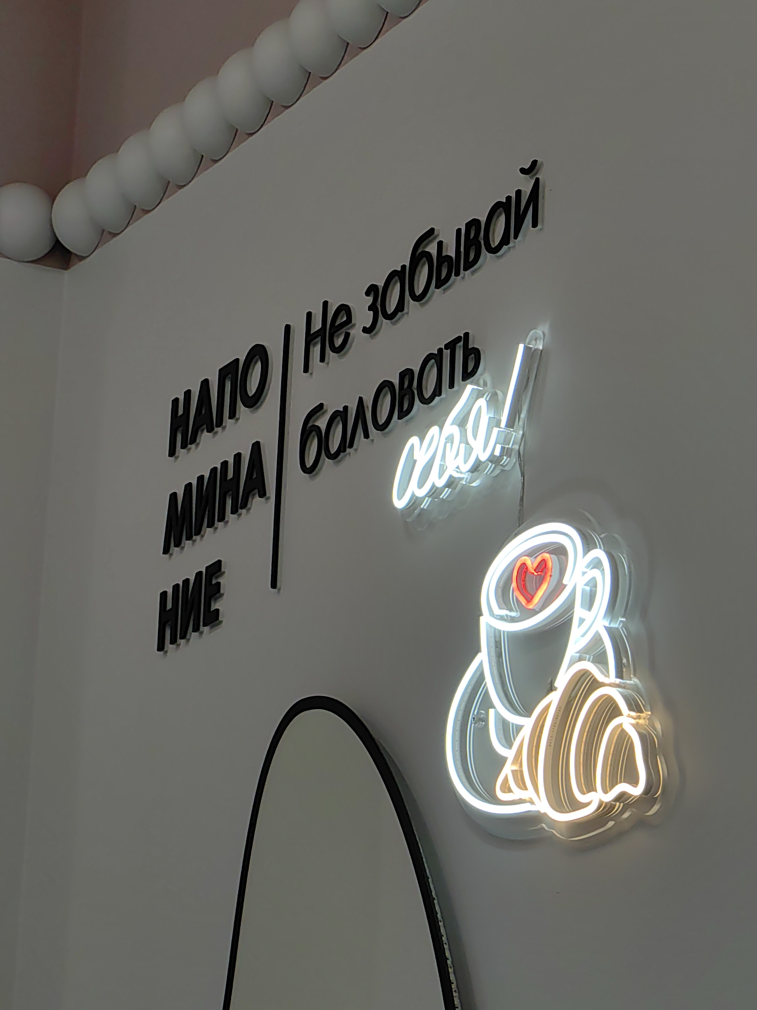An indoor scene captured in Sirius, Russia, features a light-colored wall decorated with textual messages and neon art. Along the top edge of the wall, a series of evenly spaced white spherical ornaments run horizontally, bordering a ceiling section with a soft pink tint.
The wall prominently displays a two-part message in black three-dimensional letters. On the left side, the letters vertically spell out "НАПОМИНАНИЕ" (NAPOMINANIE), which translates to "REMINDER." A slender black vertical line separates this word from the phrase on the right. The right-hand text reads "Не забывай баловать себя!" (Ne zabyvai balovat' sebya!), meaning "Don't forget to pamper yourself!" or "Don't forget to treat yourself!"
Below and to the right of the text, a luminous neon sign is affixed to the wall. It illustrates a stylized cup, within which a bright red heart glows. Adjacent to the cup, a croissant is depicted with warm, light yellow neon lighting, while the main outlines of the cup and croissant are rendered in white or light blue neon, casting a soft glow.
In the bottom left corner, a portion of a mirror with a black frame is visible. The overall ambiance suggests a welcoming and encouraging space, possibly a cafe or bakery, focusing on themes of self-care and indulgence. The image captures an interior decorative arrangement without showing any people or specific activities.