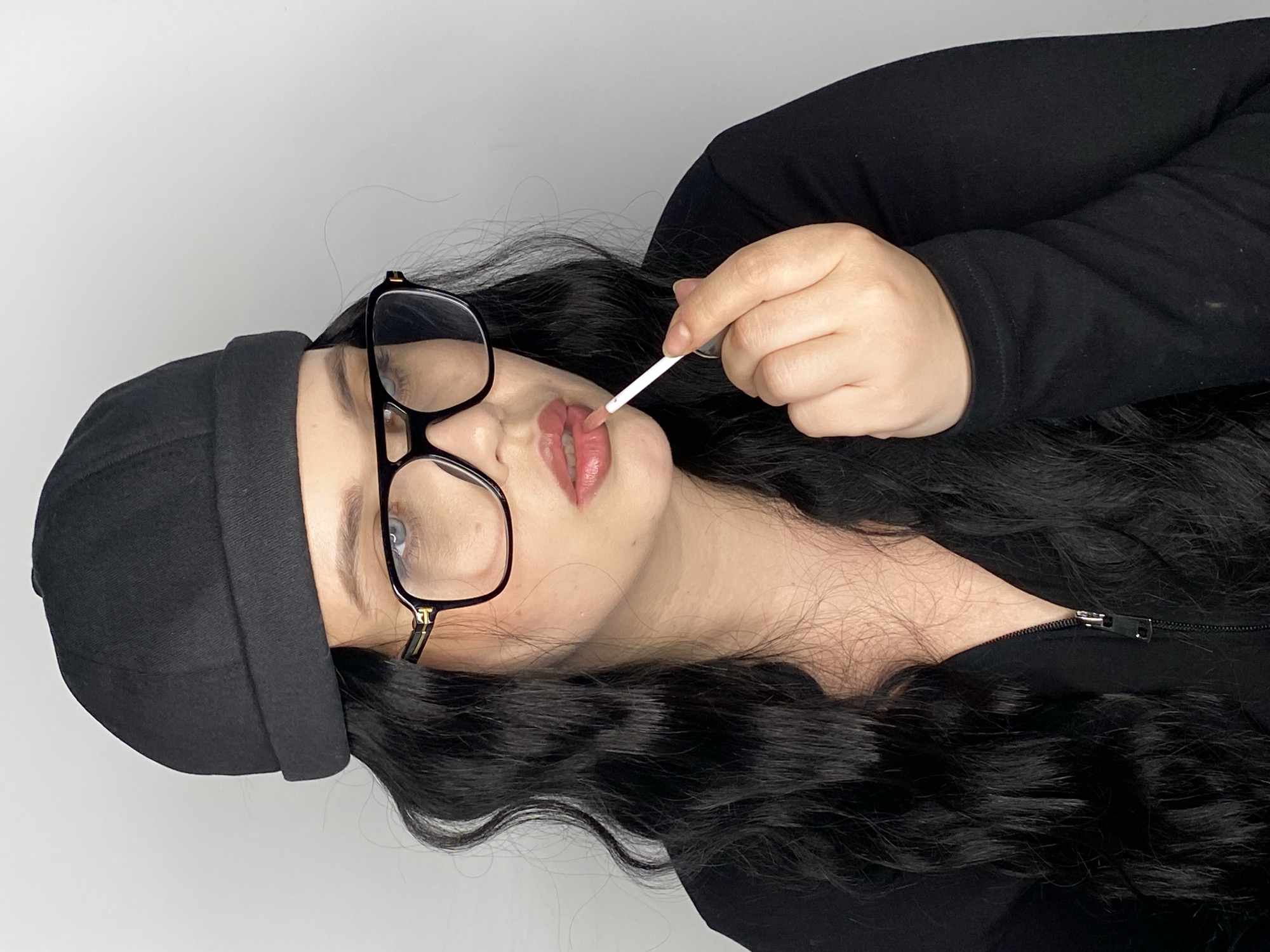 A woman is shown applying lip gloss. She is wearing a black beanie, glasses with black frames, and a black zippered jacket. Her dark hair is wavy. The background appears to be a white wall. There is no indication of the city of Chernushka, Russia in the image.