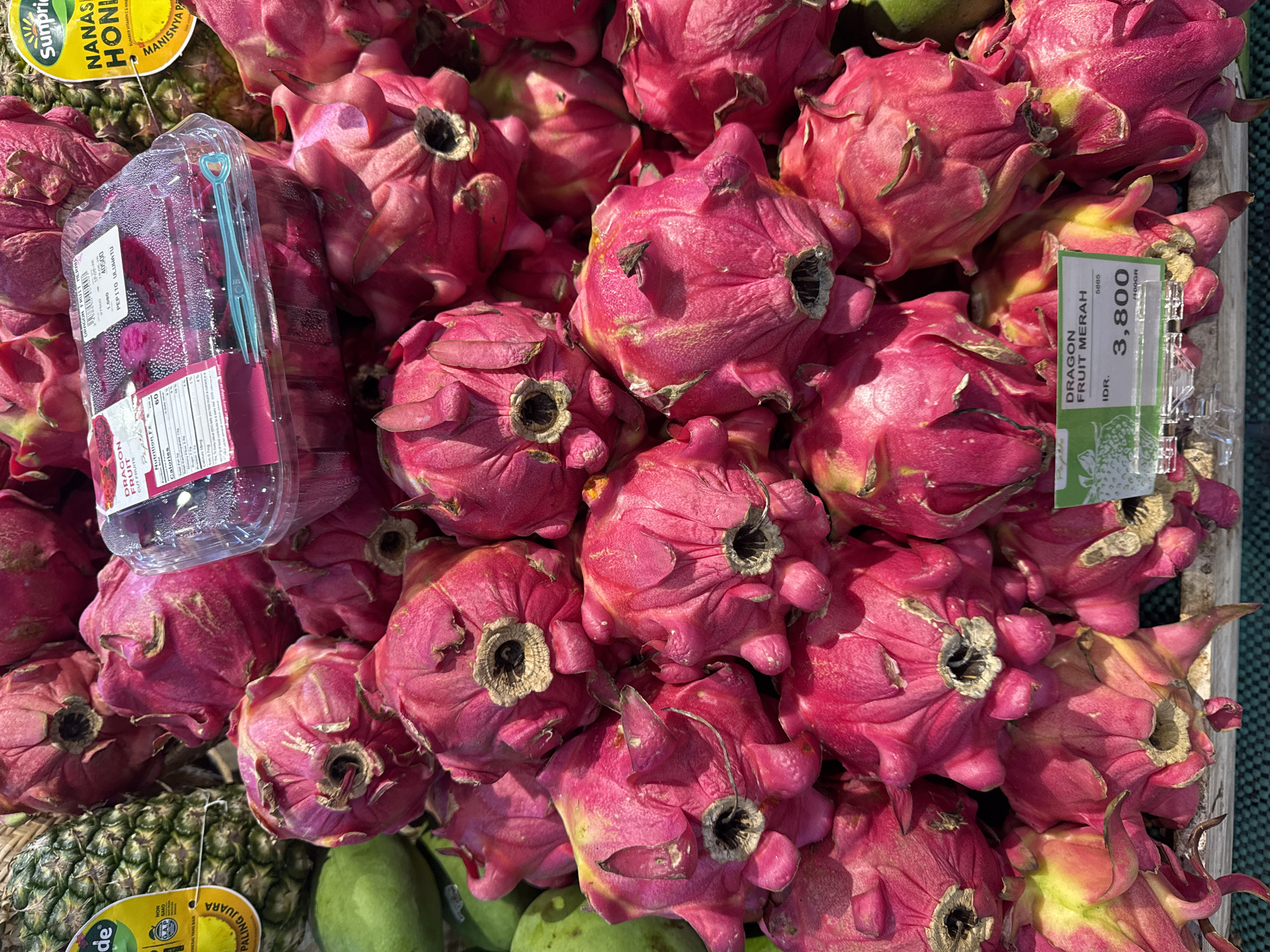 A close-up, overhead shot captures a vibrant display of dragon fruits at a market stall in Kuta Selatan, Indonesia. The foreground is dominated by a large pile of bright pink dragon fruits, their scales and stems clearly visible. A clear plastic container, partially obscured by the fruit, holds more dragon fruit. A price tag, reading "DRAGON FRUIT MERAH IDR. 3,800 /100GR," is visible on the lower right side of the container. In the background, to the left, a pineapple with its distinctive textured skin and leafy crown is partially visible, along with a green mango. The overall lighting suggests an outdoor or well-lit indoor market environment.