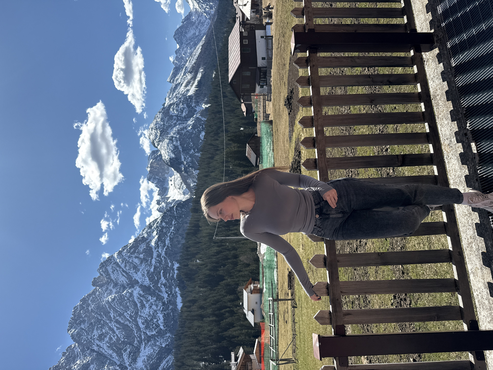 A young woman, dressed in a gray long-sleeved shirt and dark jeans, is leaning on a wooden fence in what appears to be Sappada, Italy, given the mountainous backdrop. The snow-capped mountains rise in the background under a blue sky dotted with fluffy white clouds. Houses with brown roofs can be seen nestled among the green trees and grassy fields. The woman is facing towards the mountains, resting her arm on the fence. The scene is bathed in sunlight, suggesting it is daytime. The fence has horizontal wooden slats with dark brown posts.