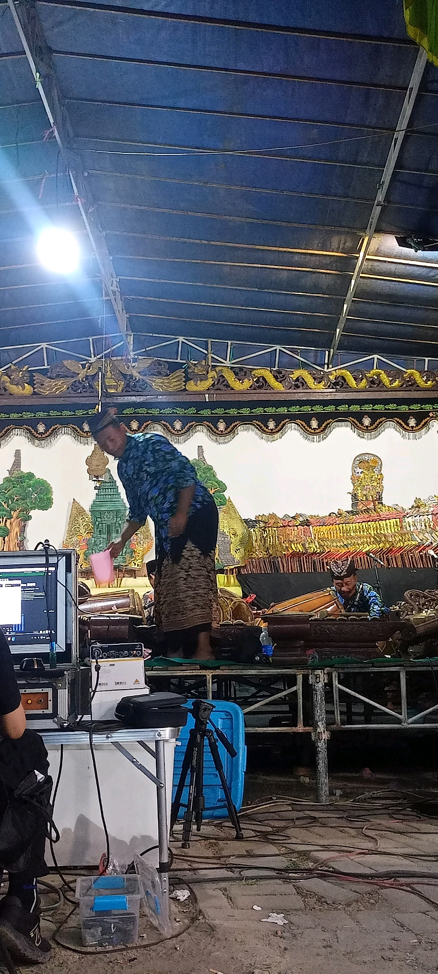 A man in a blue patterned shirt and a brown skirt stands on a raised platform in what appears to be an outdoor performance space, possibly in Gagang Kepuhsari, Indonesia. He is holding a pink plastic bucket in his left hand. In front of him, another man, wearing a blue patterned shirt and a black cap, is seated and playing a traditional Indonesian musical instrument, likely a gamelan. The background depicts a stage with elaborate decorative elements, including golden dragons and traditional Indonesian artwork. In the foreground, a technical setup is visible, with a computer monitor, audio equipment, and a tripod with a camera, suggesting a live recording or broadcast of the performance. The overall environment appears to be a preparation or performance area for a cultural event.
