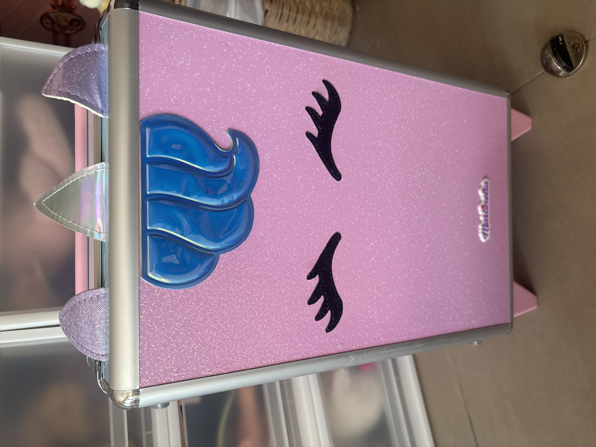 A pink glitter-covered suitcase designed to resemble a unicorn sits on a brown floor in front of a set of clear plastic drawers. The suitcase has blue, wavy hair molded onto the front, with black eyelash stickers above it. Two iridescent unicorn ears are attached to the top edge of the suitcase. A small, silver charm is attached to the suitcase by a thin string. The background suggests an indoor environment, possibly a bedroom or storage area. There is no indication of the suitcase's location being in Dubai, United Arab Emirates.