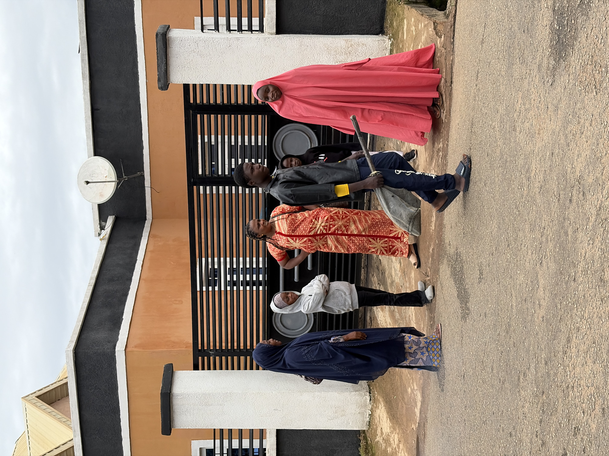 In the city of Jos, Nigeria, a group of six individuals stands near a modern residential building. The building features a dark grey band along its top with white trim, resting above an orange facade. A black metal gate with horizontal slats separates the property from the street.
On the left, a woman wearing a dark blue hijab and a patterned dress stands facing right, her left hand resting on her hip. To her right, a young person in a grey hooded sweatshirt and dark pants stands with arms crossed. Next to them, a woman in a vibrant red and gold patterned dress, her hair visible, also stands with her hands on her hips, looking to her left.
In the foreground, a young man wearing a black leather-like jacket over a yellow shirt and dark pants walks from right to left. He is carrying a grey bag or bundle on a stick, with the stick held in his right hand. To his right, a person entirely draped in a bright pink garment, covering them from head to toe, stands facing forward. Partially visible behind the young man and to his left is another young person in a black hooded garment.
The ground is paved with asphalt, appearing slightly wet or discolored in places. The sky is overcast with grey clouds. The overall scene depicts a moment of pause or waiting on a street in Jos.