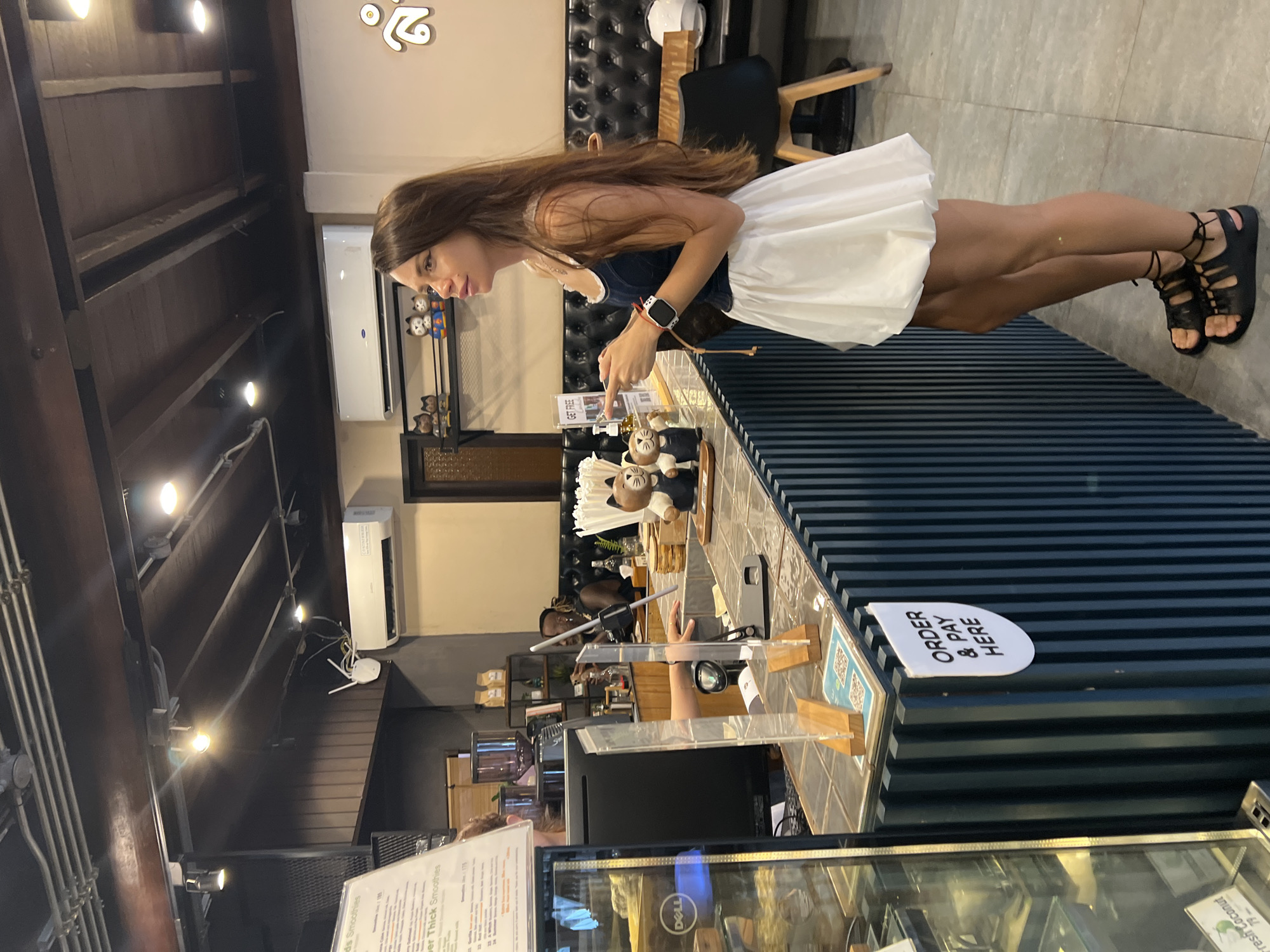 The image captures the interior of a commercial establishment, possibly a café, in Baan Lamai, Thailand. A woman with long brown hair, wearing a dark top, white skirt, and black sandals, stands at the counter, examining items displayed on the countertop. The counter has a horizontal stripe design with a sign reading "Order & Pay Here". A transparent barrier is present on the counter. In the background, shelves holding various items are visible. The lighting fixtures and pipes are visible near the ceiling. An air conditioning unit is on the wall. A white object on a chair is in the background to the right. An illuminated sign is visible on the upper wall, and text is partially legible on a menu in the lower left corner, with the words "Smoothies" and "Fresh Coconut". A Dell monitor is visible below the countertop glass.