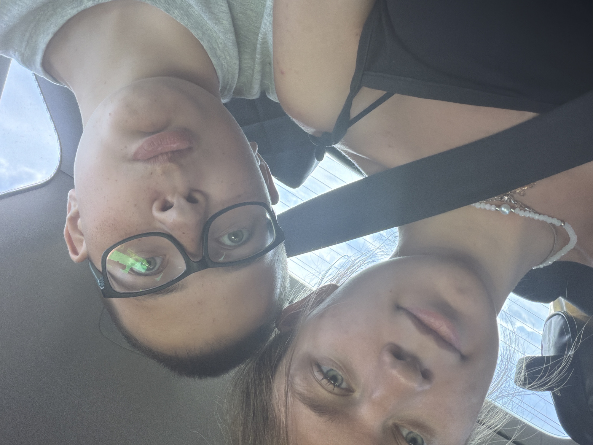 This is a close-up, low-angle selfie taken inside a vehicle. Two young individuals are visible, with the camera angled upwards to capture their faces. The person in the foreground, on the left, has light brown hair and fair skin with freckles. They are looking directly at the camera with green eyes. They are wearing a black tank top and a beaded necklace. Behind them, on the right, is another person wearing glasses with black frames. This individual has fair skin, also with freckles, and is wearing a light grey t-shirt. Their eyes are visible through the glasses, and their lips are slightly parted. A black seatbelt is prominently visible across the chest of the person in the foreground. The background shows a bright sky and a portion of the vehicle's interior, including the ceiling and window. No specific location within the city of Uziukovo, Russia, can be determined from the image.