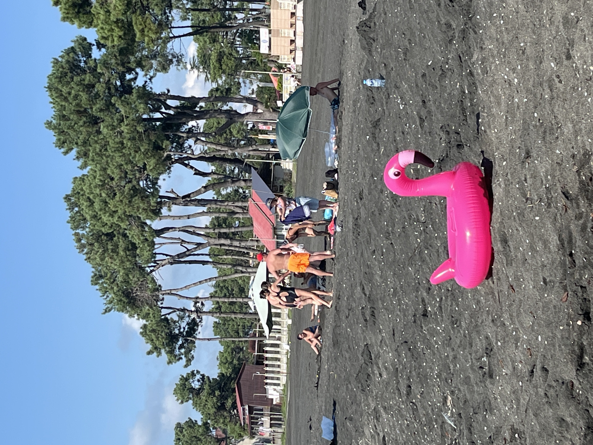 A group of people are on a black sand beach under a clear blue sky with scattered clouds. In the foreground, a bright pink inflatable flamingo lies on the sand. In the midground, several people are standing and sitting. Some are in swimwear, with one person wearing orange shorts and another a black swimsuit. An umbrella provides shade for a person lying on the sand. Several buildings and tall trees are visible in the background, suggesting a coastal town or resort area, consistent with Ureki, Georgia. The overall scene depicts a relaxed beach day.