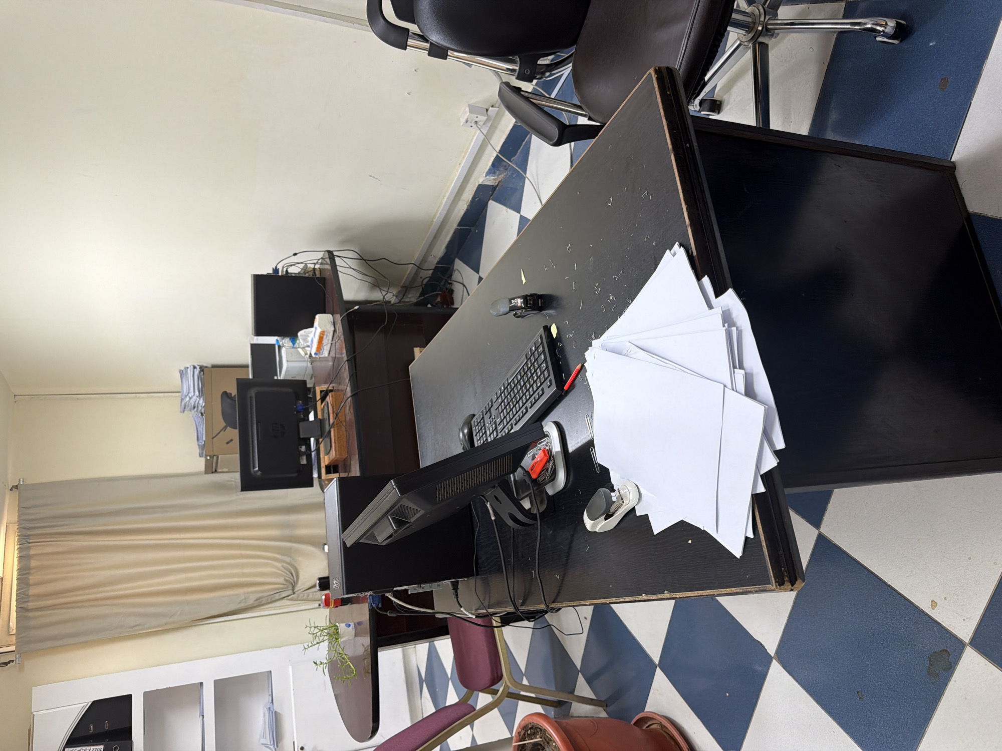 The image shows a cluttered office space in Sharm El Sheikh, Egypt. In the foreground, a large, dark desk is covered with various office supplies, including a computer monitor, keyboard, mouse, stapler, and stacks of paper. A red pen and a red USB drive are also visible on the desk. To the left, a burgundy chair with a metallic frame is positioned near the desk. A potted plant sits on a smaller, curved desk behind the main desk. In the background, a tall white shelf unit with folders and papers is visible against a wall with a beige curtain. Further back, another desk holds two computer monitors and a black computer tower. Cardboard boxes are stacked on a shelf in the background. The floor is tiled with a blue and white checkerboard pattern. The overall impression is one of a functional but somewhat disorganized workspace.