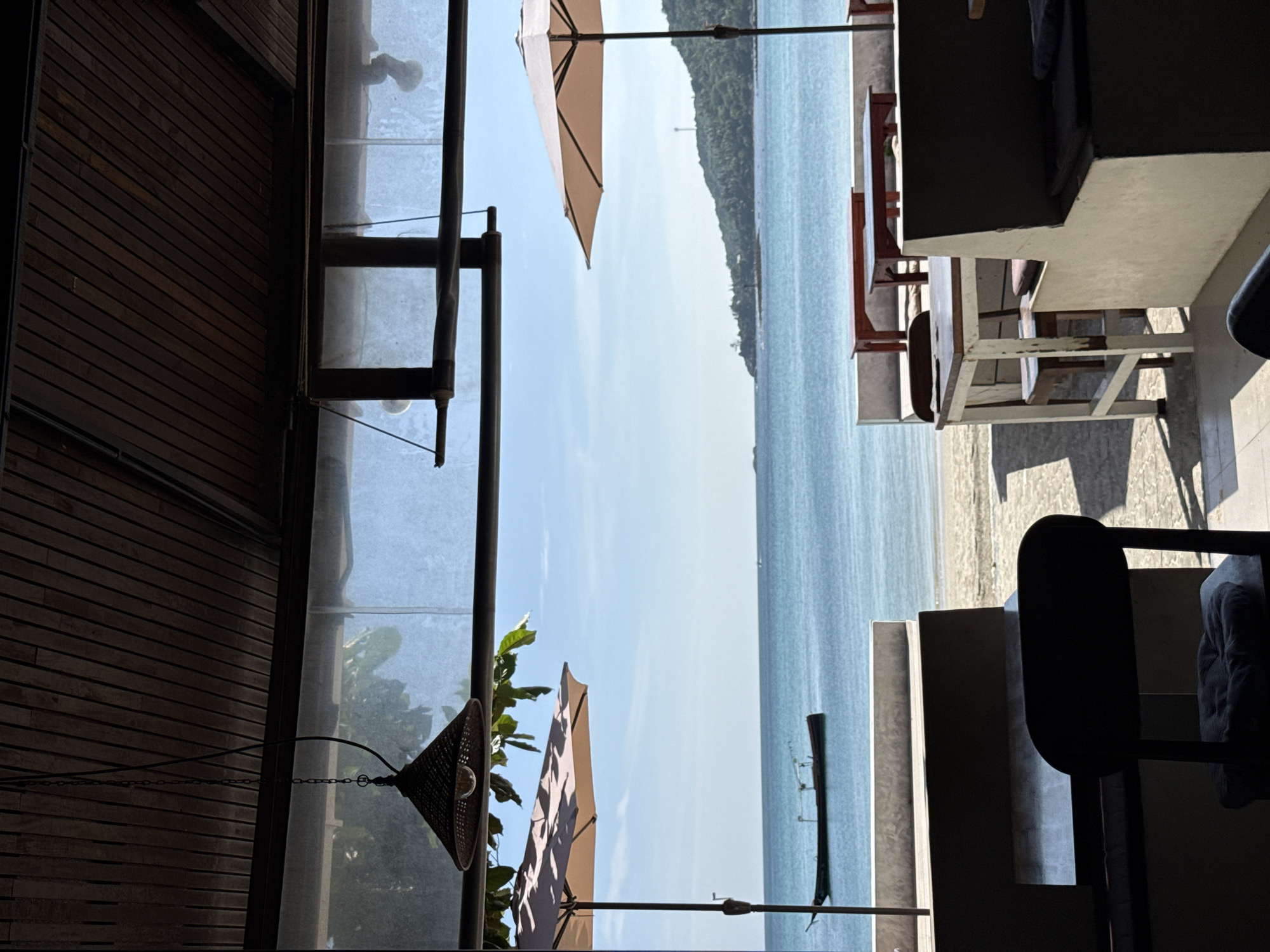 The image depicts an outdoor seating area overlooking a tranquil beach and turquoise ocean in Ko Pha-ngan District, Thailand. In the foreground, several tables and chairs are arranged, some with red wooden bases and others with white frames. A dark chair with a cushioned seat is prominently positioned in the lower left corner. The seating area is partially covered by a wooden ceiling and features two beige umbrellas, one on the left and another on the right.
Beyond the seating area, a sandy beach stretches towards the water, which is calm and reflects the clear blue sky. A narrow, dark-colored boat with an elevated structure is moored in the shallow water. In the distance, a lush, green hillside slopes down to the sea, with a small tower visible near its peak. The overall atmosphere is serene and inviting, suggesting a peaceful seaside dining or relaxation spot. The time of day appears to be daytime, with bright sunlight casting shadows on the ground.