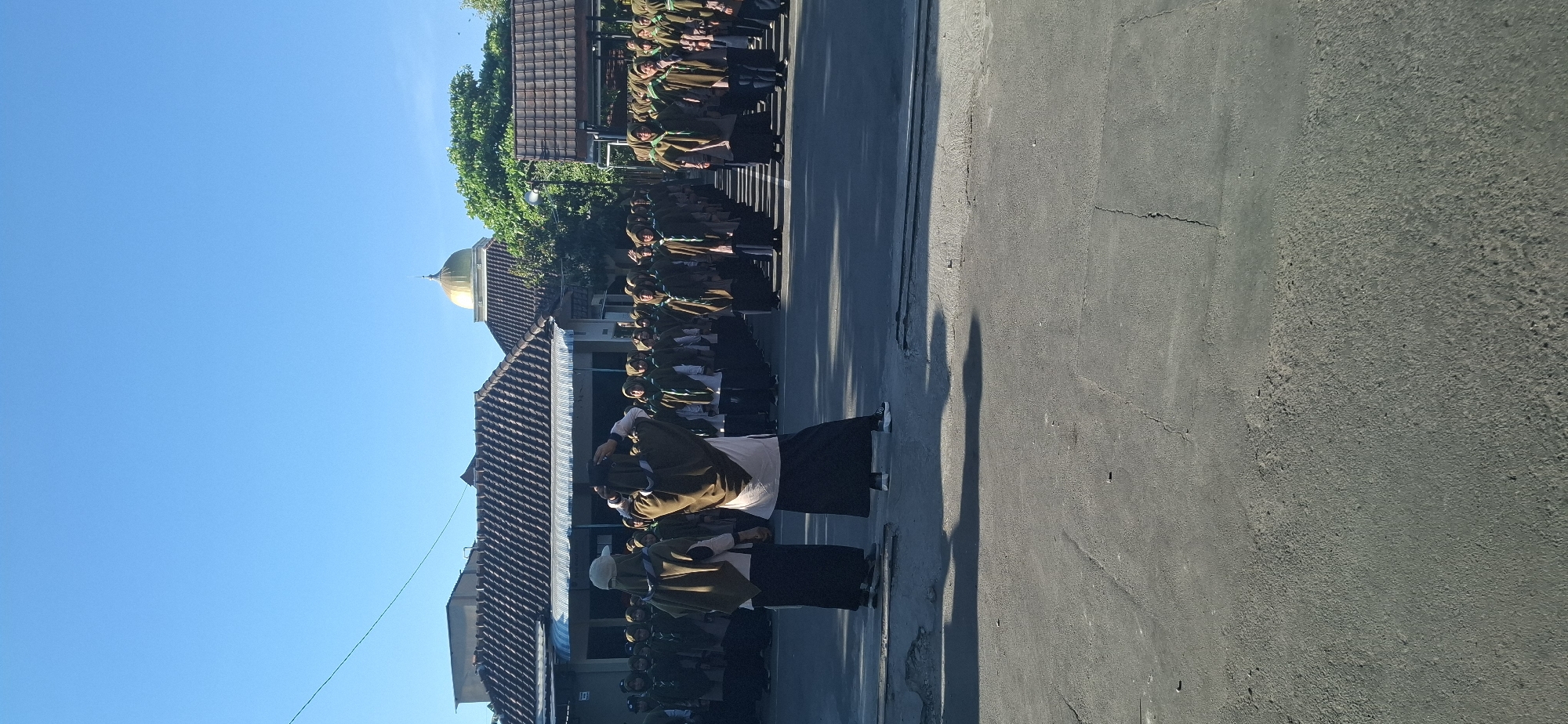 In Wonorejo, Indonesia, a group of people, predominantly women in hijabs and long skirts, are gathered outdoors in formation. They appear to be participating in an organized activity or event, possibly a school assembly or a scouting activity, given their matching brown vests and green accents. Two individuals in the foreground are facing away from the camera, with one adjusting her hijab. Behind them, rows of participants stand in neat lines. The background features buildings with tiled roofs, one of which has a distinctive golden dome, suggesting it might be a mosque or religious building. Trees and a clear blue sky complete the outdoor scene, with a paved road in the foreground. The overall impression is one of an orderly gathering in a communal or educational setting.