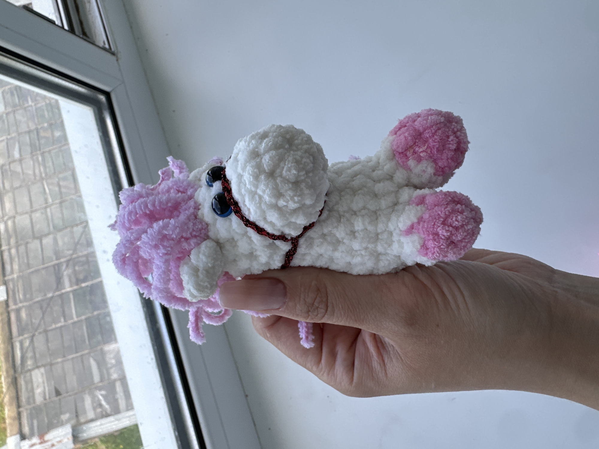 A hand with a light pink nail polish holds a white and pink crocheted unicorn toy in front of a window. The unicorn has blue plastic eyes, a red beaded halter, and a pink mane. The window behind it shows a blurry view of what appears to be a greenhouse or industrial building with a grid pattern and some greenery. The scene is lit from the outside, casting a soft light on the toy and hand. The background is a plain white surface. The image does not provide any information about the specific location in Yaroslavka, Russia.