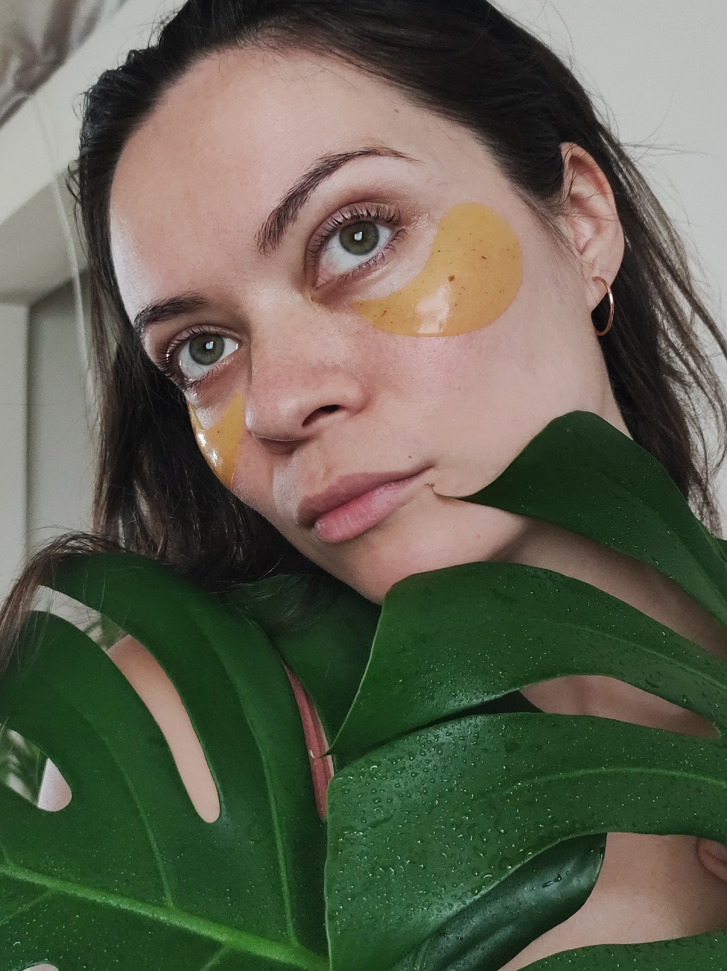 A woman with dark hair and green eyes looks upwards while wearing gold under-eye patches. She is partially obscured by a large, green Monstera leaf covered in water droplets. She has a fair complexion and is wearing gold hoop earrings. The background appears to be a plain, light-colored wall with a hint of a doorway or corner. This image has no visible relation to the location Фоки, Russia.