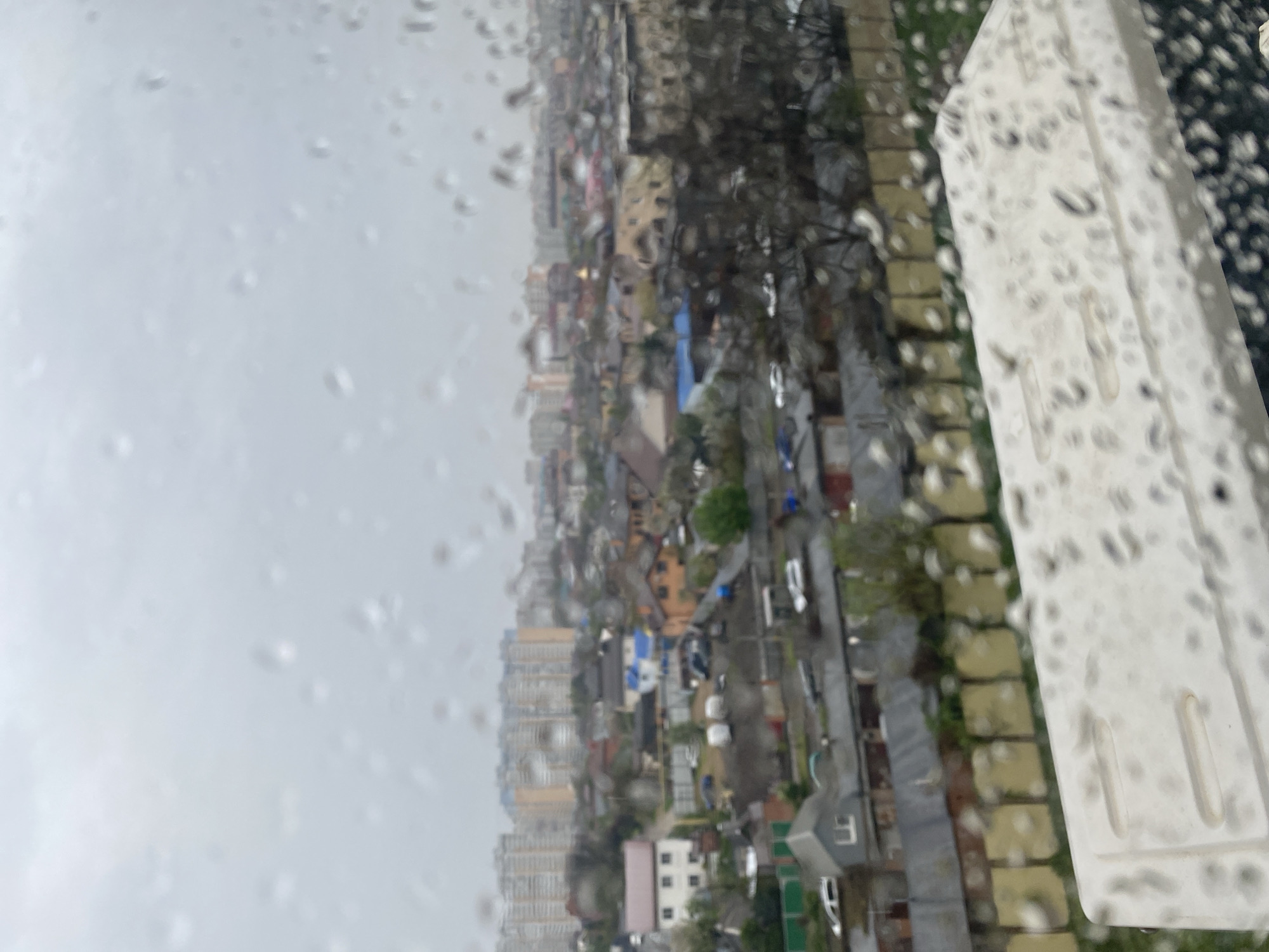 The image shows a cityscape under rainy conditions. Raindrops are visible on the foreground surface, likely a window. The city features a mix of high-rise apartment buildings and smaller residential houses with varying roof colors. Streets are visible, with parked cars scattered along them. There are trees among the buildings and along the streets. A part of what appears to be a balcony or window frame is visible in the right foreground, covered with raindrops. The scene has an overcast and wet atmosphere due to the rain.