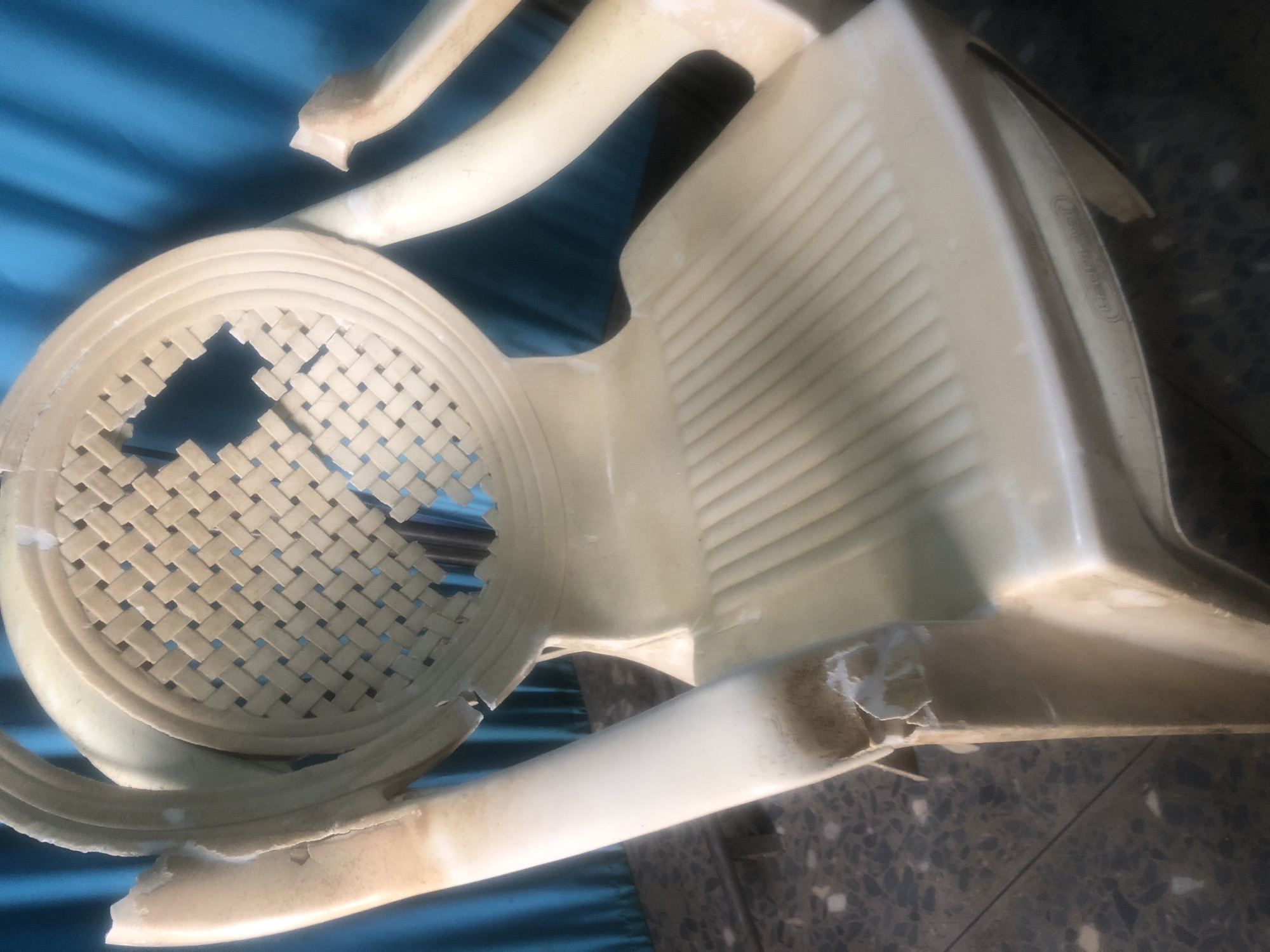 The image shows a damaged, off-white plastic chair with a woven back that has several holes. The chair is outdoors, with a blue curtain in the background and a speckled floor. The chair appears to be in a state of disrepair, with cracked and peeling plastic on the arms and back. There are no people or animals visible in the image. The setting is likely a residential area or a public space in Jalingo, Nigeria, given the context provided.