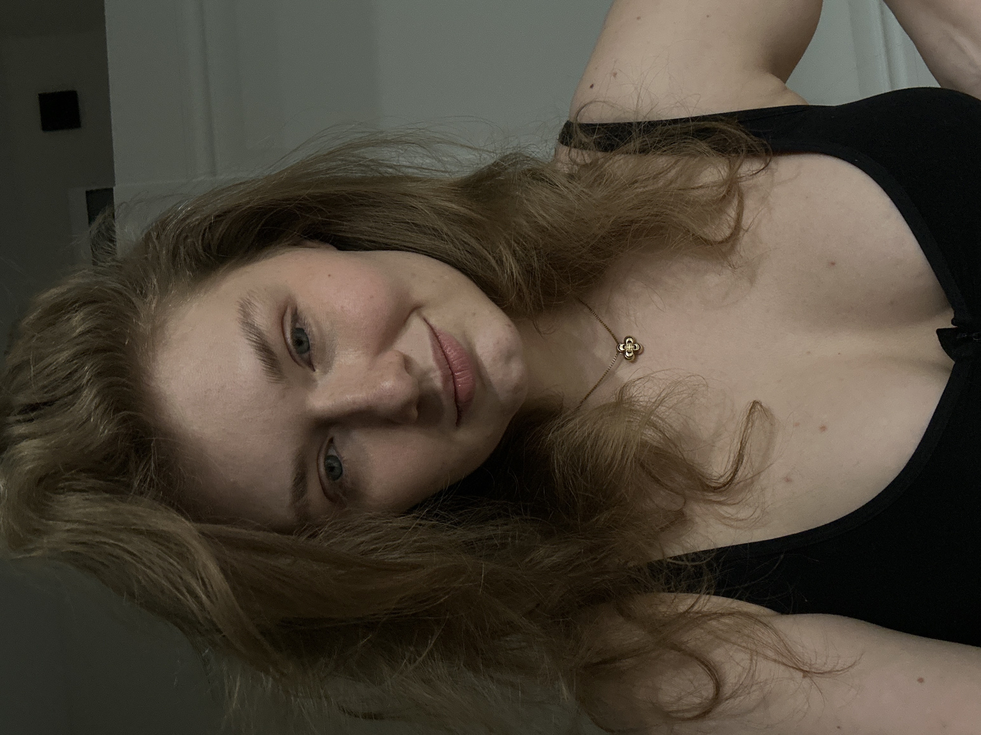 A woman with fair skin and light brown hair is pictured indoors. She is wearing a black top and a gold necklace with a flower-shaped pendant. The background includes a wall and a dark square shape, possibly a light switch. The photo appears to be taken at an angle, tilting the woman's face. There is no identifiable information that places the image in Kazan, Russia.