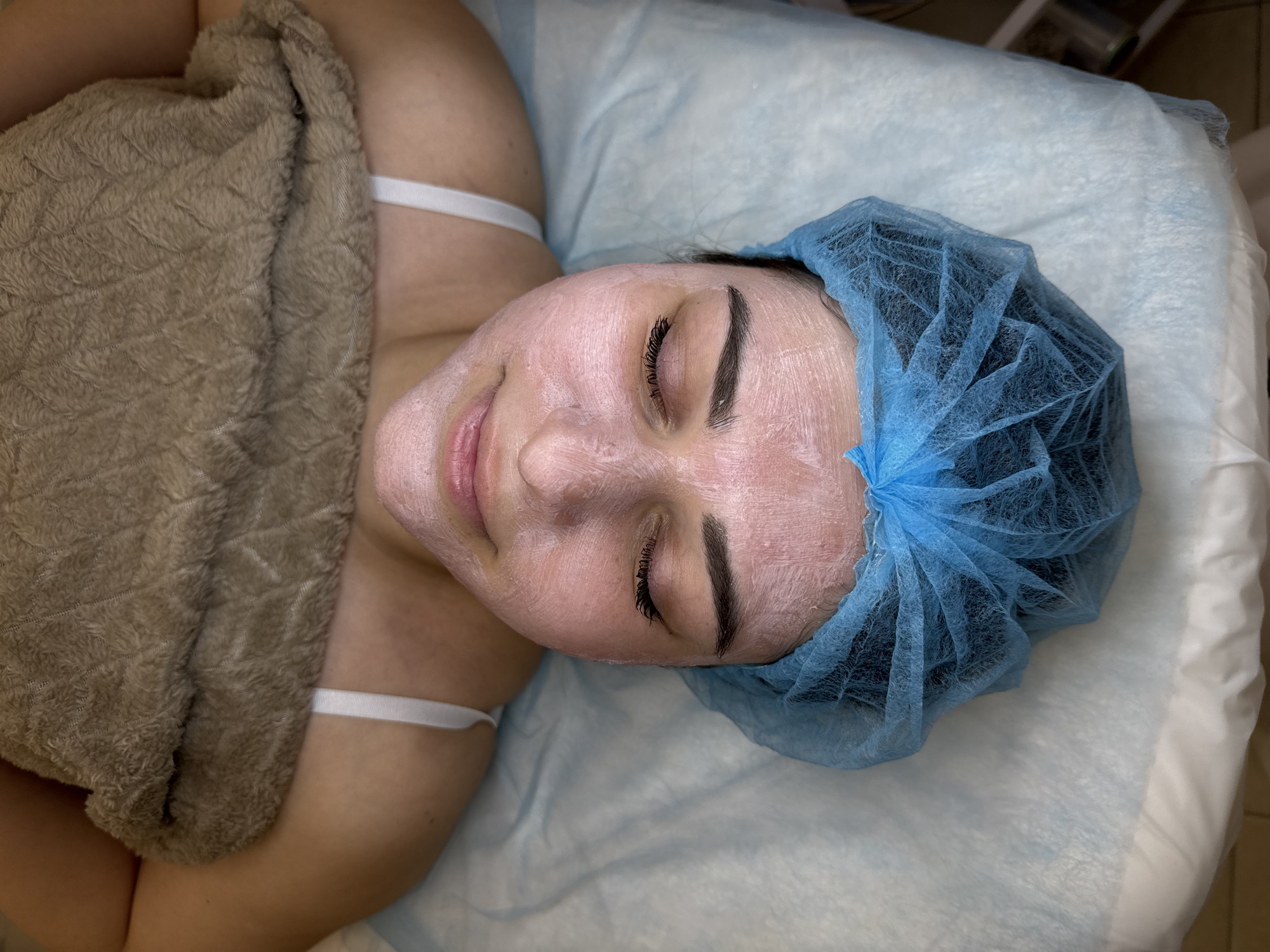 An overhead shot shows a person lying down, seemingly receiving a facial treatment. The individual, likely female, has their eyes closed and a white or pinkish mask applied to their entire face. A blue, disposable bouffant cap covers their hair. They are lying on a surface covered with a light blue disposable sheet, commonly found in spa or medical settings. A textured, brown blanket or towel covers their torso, revealing a white bra strap on their left shoulder. The person's lips are slightly parted in a faint smile, suggesting a state of relaxation. The background is mostly out of focus, with some indistinct shapes visible in the upper right corner. There is no visible text.