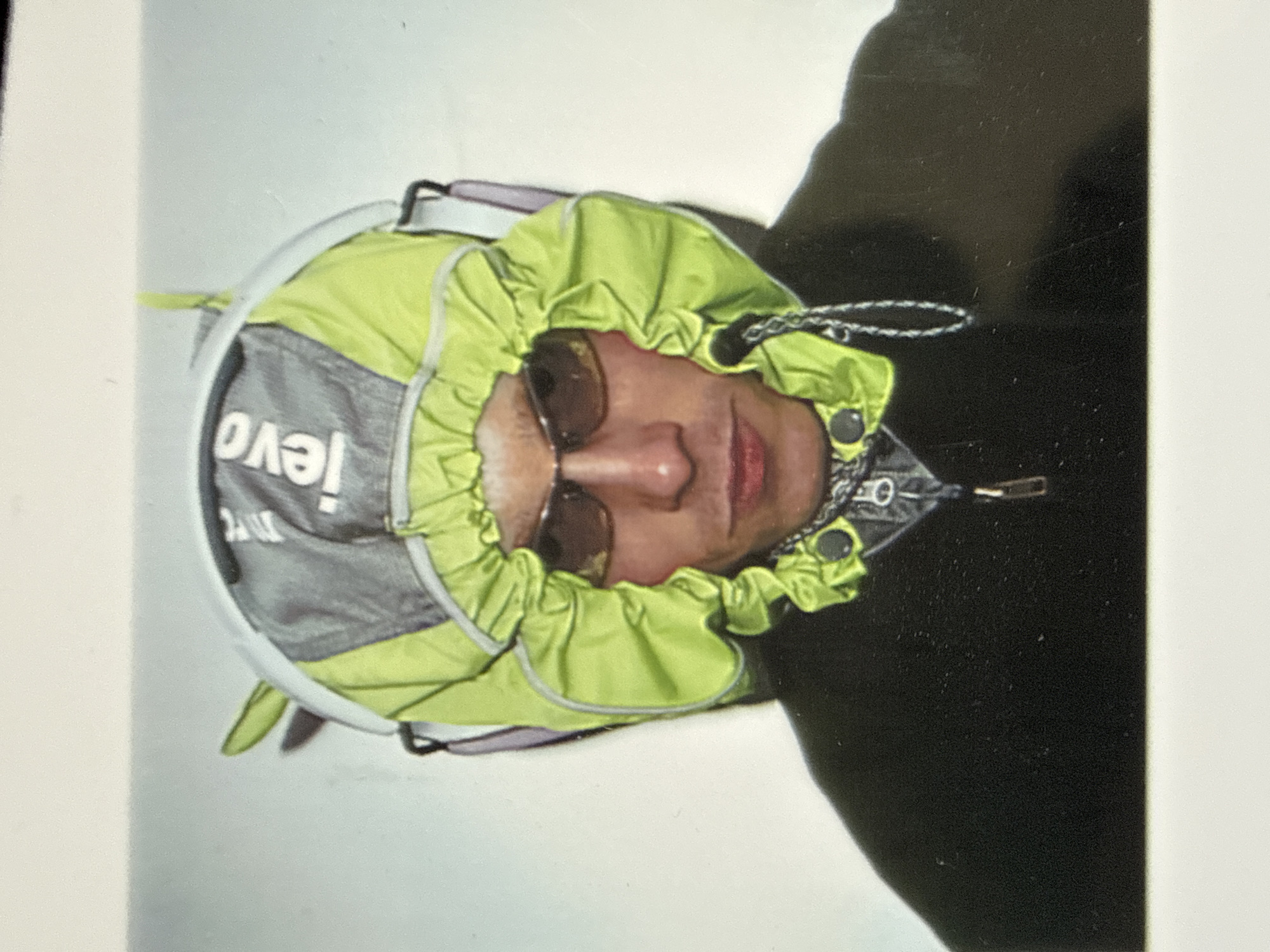 A person wearing sunglasses is pictured with the hood of a gray and bright green backpack cinched around their face. The backpack has the word "levo" written on it. The person is wearing a black jacket with a zipper. The background is mostly white. The location is not identifiable as being in London, United Kingdom.