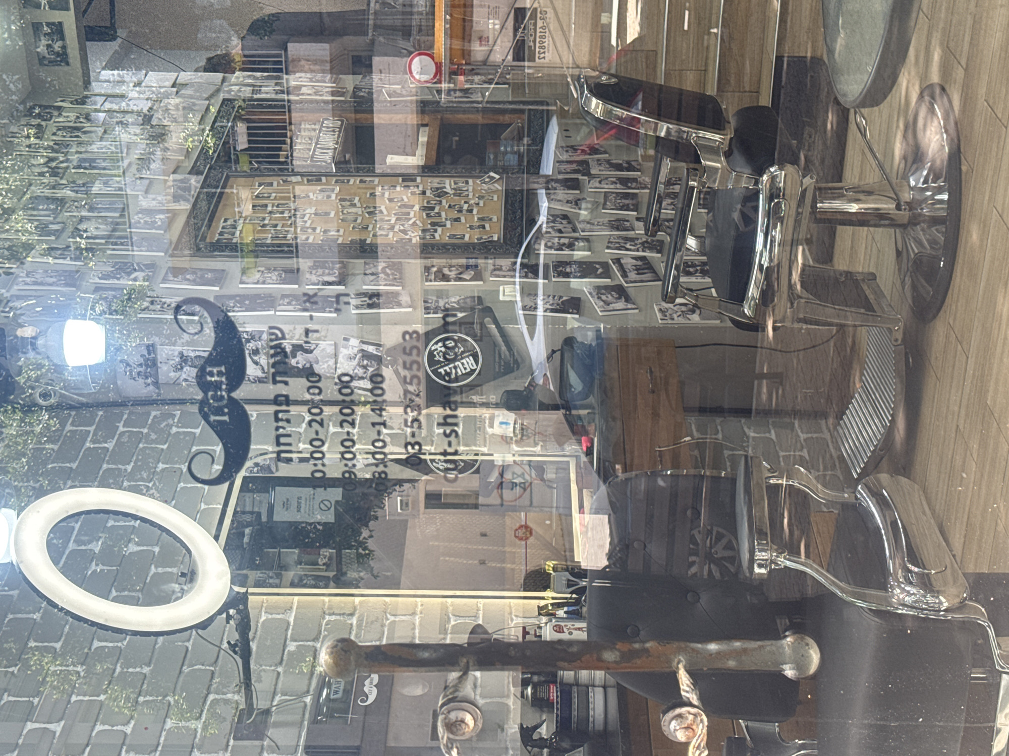 The image depicts the interior of a barbershop, likely located in Tel Aviv, Israel. The shop features classic barber chairs, one with a ring light behind it. The front window is covered with numerous black and white photographs, and a sign with a mustache logo. The sign also displays the shop's name, "IGH," opening hours, and phone number: 03-5375553. There is a reflected view of a city street.