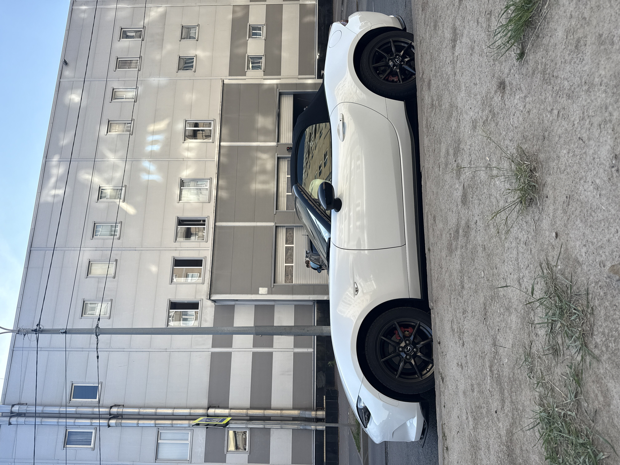 The image depicts a white convertible Mazda MX-5 parked on a city street in Saint Petersburg, Russia. The car is positioned parallel to a sidewalk made of concrete with some sparse grass growing through it. Behind the car is a large, modern building with a gray and white striped facade. The building has numerous windows arranged in horizontal rows, and a section of the lower facade appears to be a garage or entrance with rolling doors. A street lamp post stands in front of the building. There are two individuals in the background near the garage doors. The sky is a clear blue with some diffused sunlight creating reflections on the building's surface.
