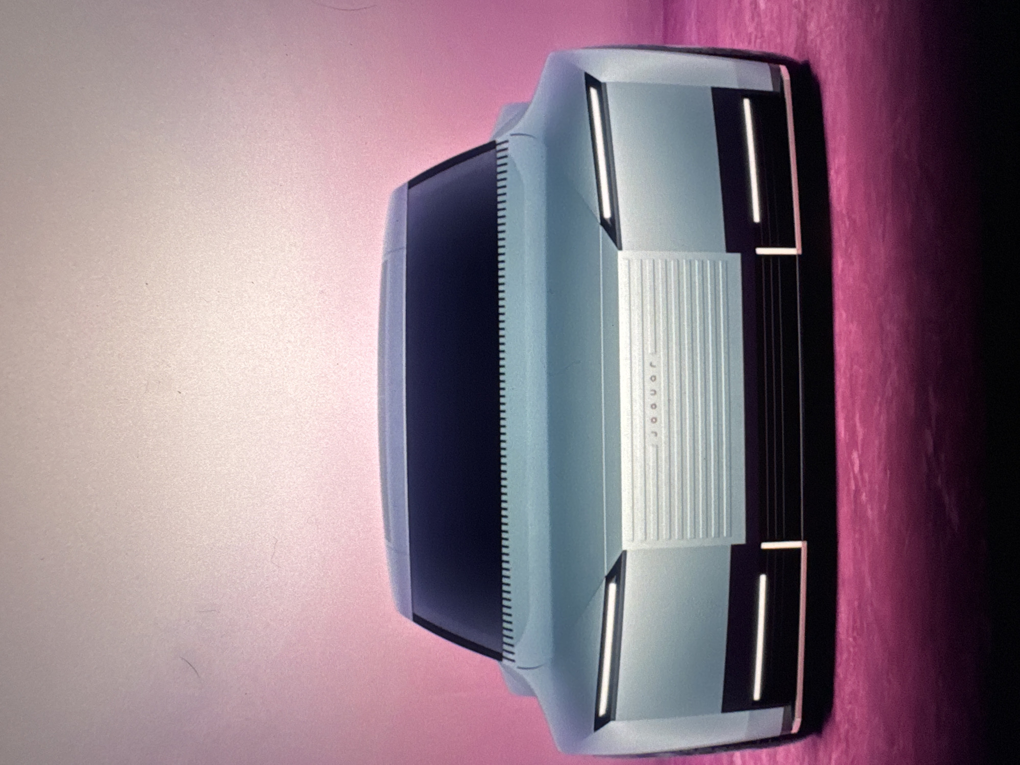 The image displays the front view of a futuristic, light blue concept car with a black grille and thin, illuminated headlights. The car is positioned against a pinkish-purple background. The car's design is sleek and minimalist, featuring sharp lines and a flat front. A subtle "jaguar" logo is visible on the grille. The overall impression is one of a high-tech automotive design study. The image does not provide any information about the location in Sant'Angelo di Piove di Sacco, Italy.