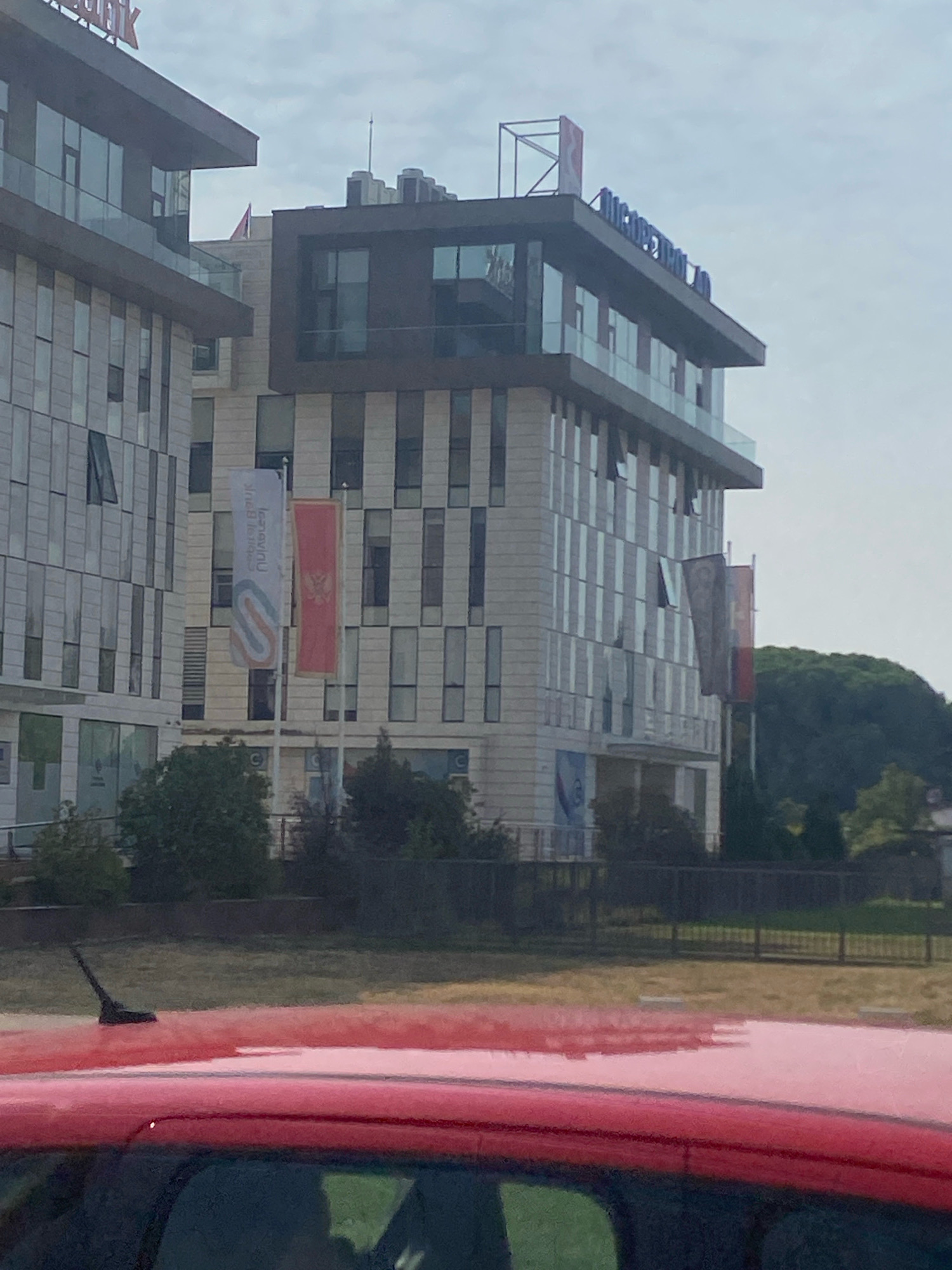 The image shows a modern office building complex located in Podgorica, Montenegro. The building is characterized by its glass facade and multi-story design, with some sections featuring dark cladding. Two flags are visible on flagpoles in front of the building: one is a vertical white banner with a blue and orange logo, and the other is the national flag of Montenegro, featuring a red background with a gold border and a gold coat of arms. The building is situated in an urban environment with a road and some dry grass in the foreground, partially obscuring a red car's roof and antenna. Trees and a hill covered with foliage are visible in the background, suggesting a location on the outskirts of the city or in a greener area of Podgorica. The sky is overcast. The building appears to be a commercial or corporate establishment.