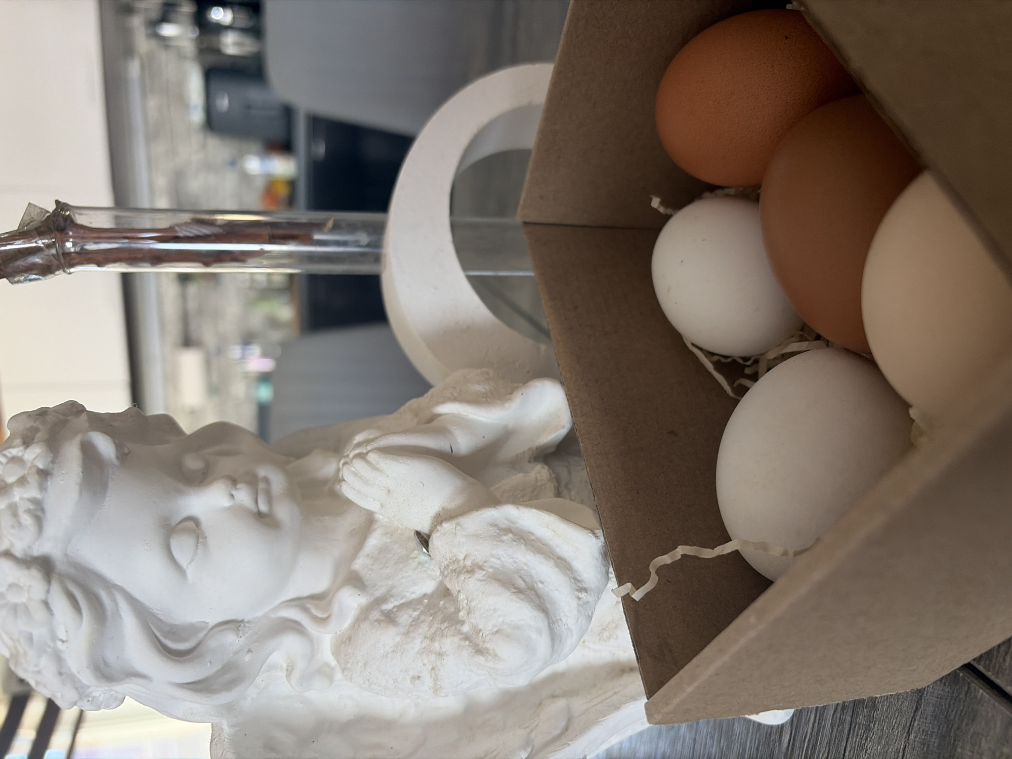 The image shows a still life arrangement. A white plaster figurine of a child with hands clasped in prayer is positioned next to a cardboard box containing several eggs of varying colors (brown and white). A glass tube extends horizontally near the top of the figurine. The image appears to be taken indoors.