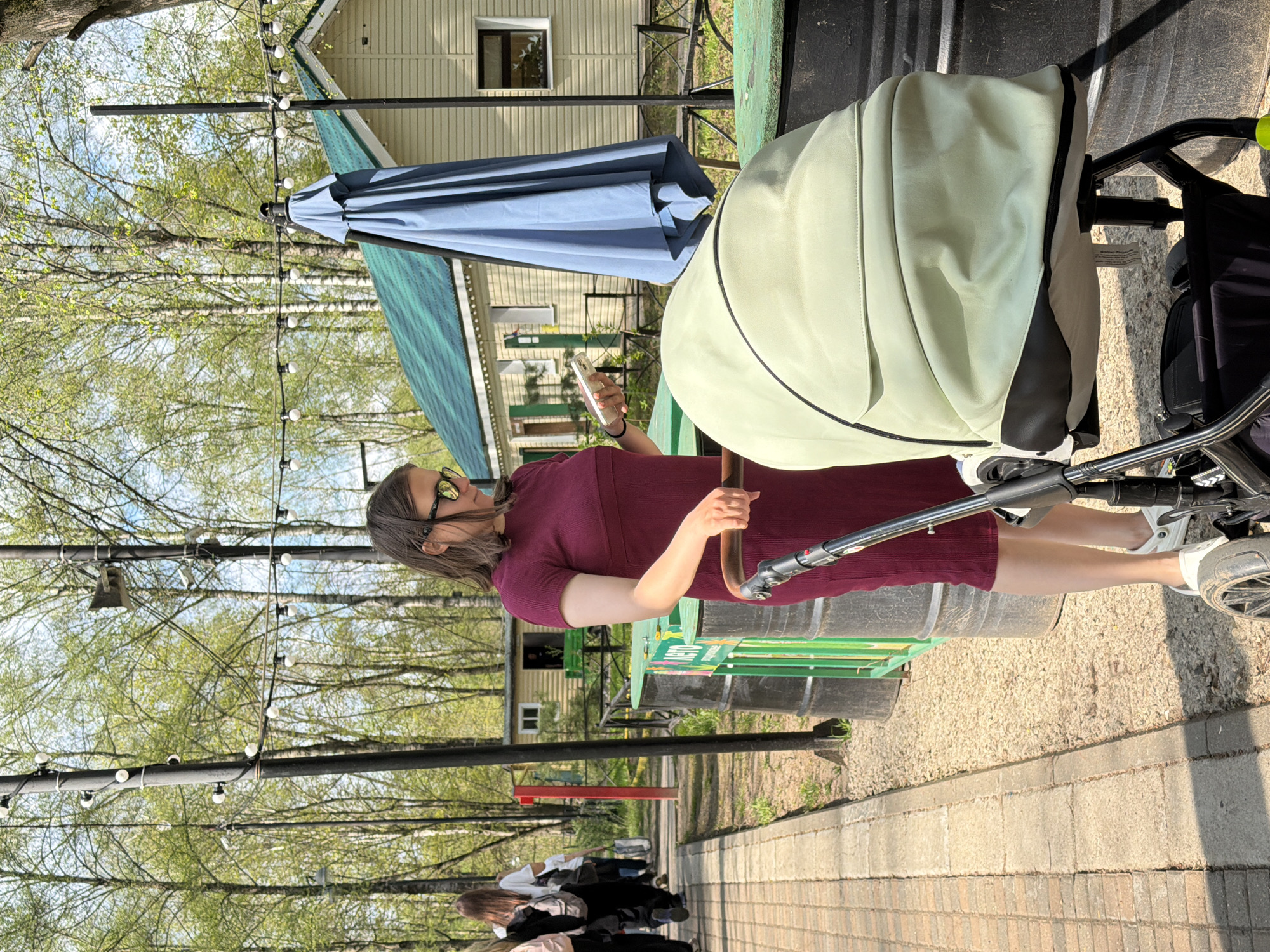 The image depicts a woman standing next to a baby stroller on a pathway. The woman is wearing a maroon dress and sunglasses, and she is holding a smartphone in her hand. The stroller is light green with a black trim. The pathway is paved with brick and stone, and it is lined with trees. There are people walking in the background. The scene takes place in what appears to be a park or outdoor recreational area, potentially within the city of Klin, Russia, as inferred from context.